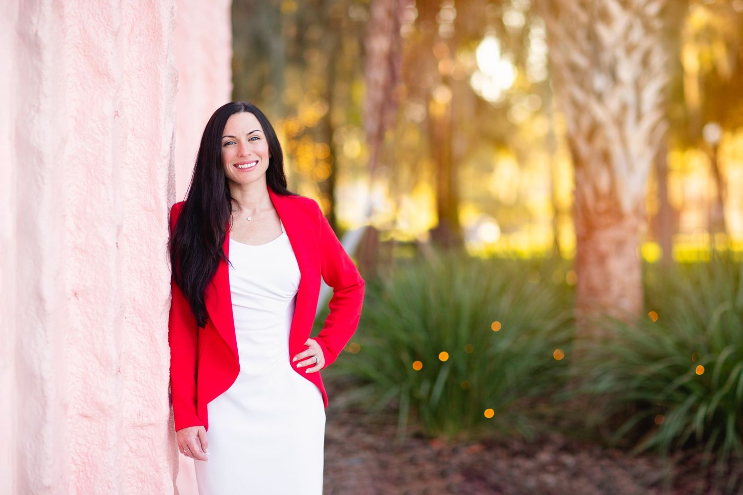 Professional portrait featuring red blazer and white dress against tropical outdoor background.