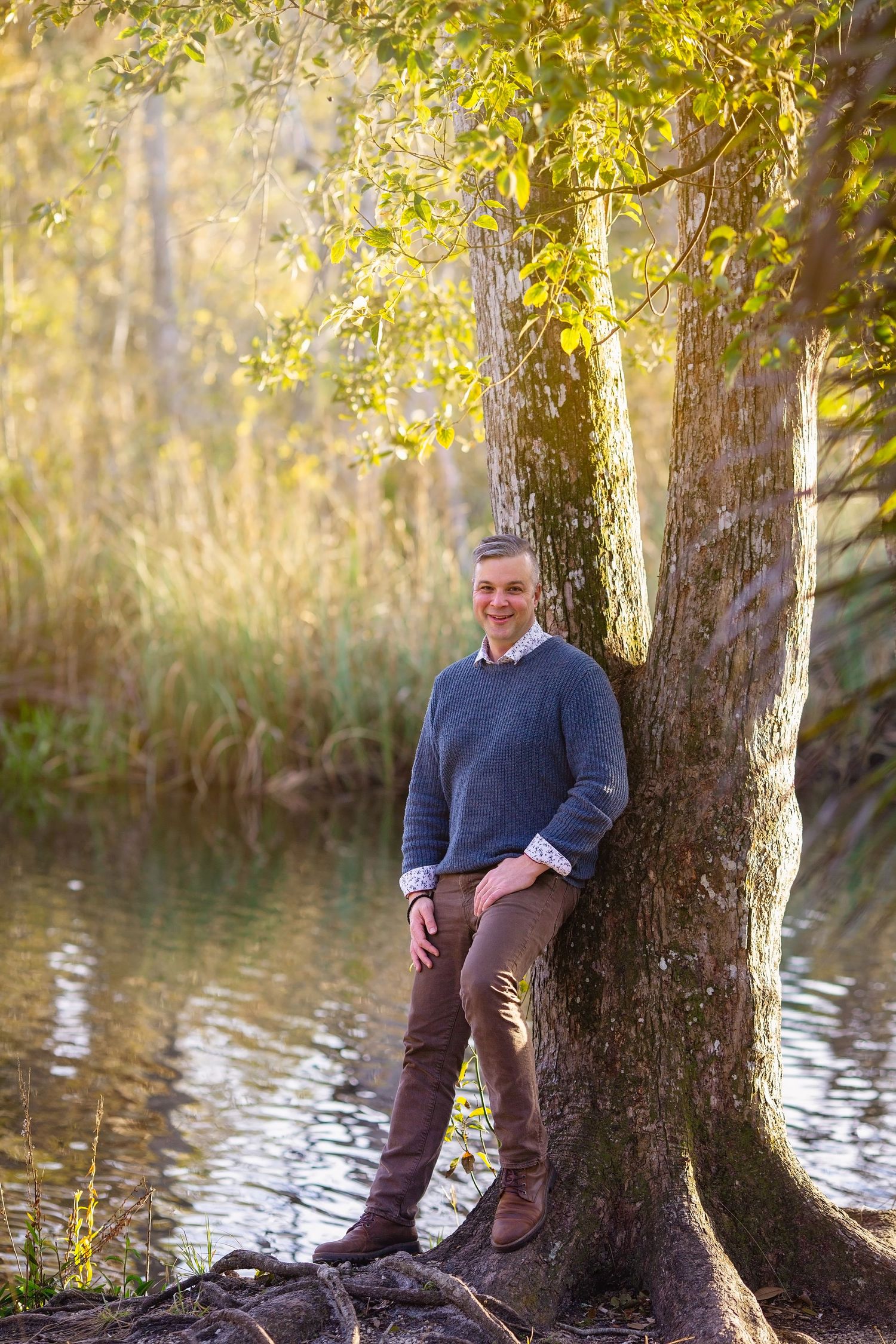 Casual portrait of person leaning against tree trunk by water in afternoon sunlight.