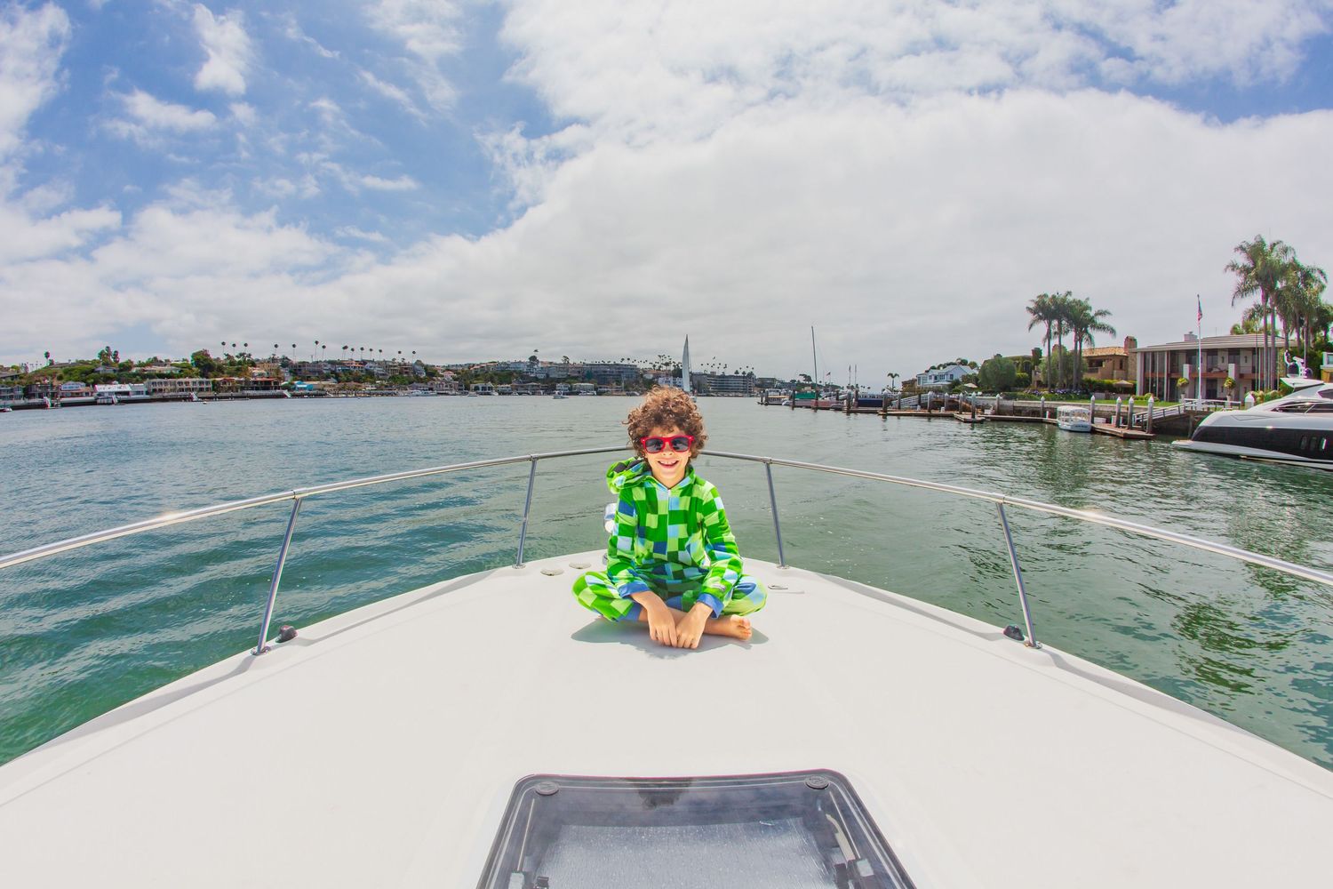 Child in green outfit sitting on white boat deck with waterfront view.