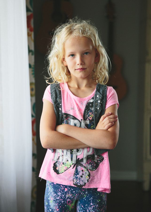 Child in pink butterfly shirt and floral vest standing with crossed arms indoors.
