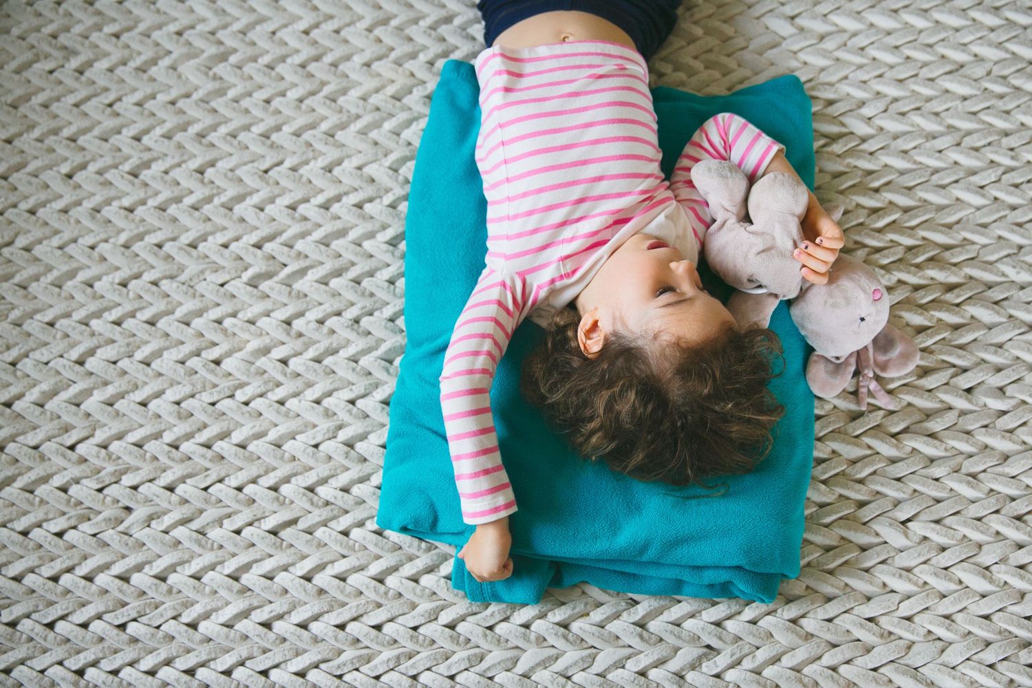 Small child lying on a turquoise cushion on a textured carpet wearing pink striped clothing.