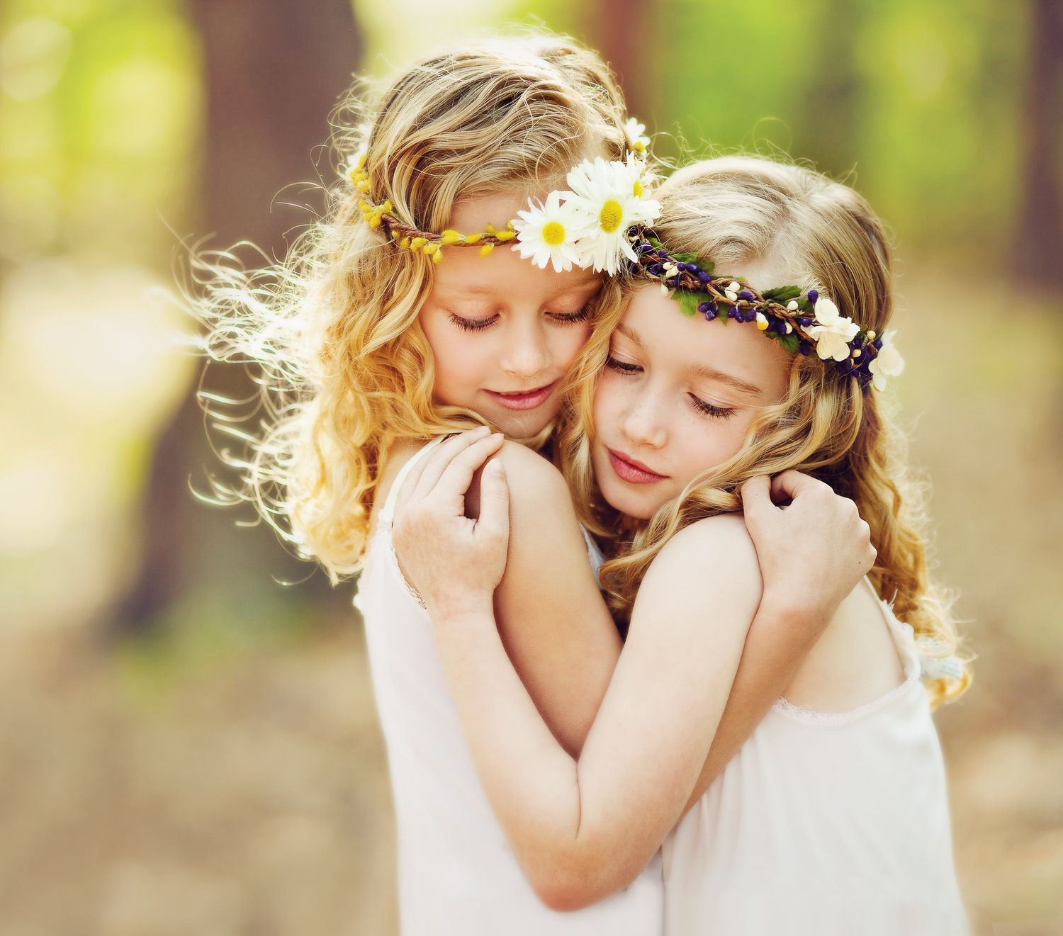 Two children wearing white dresses and flower crowns embracing in soft sunlight.