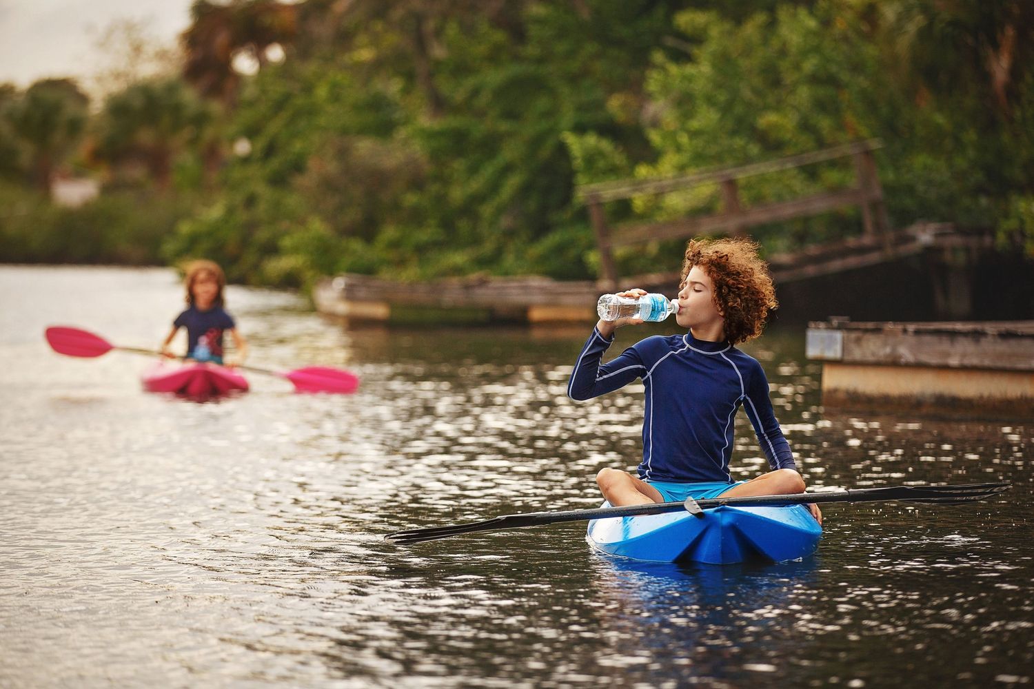 Someone drinking water while kayaking on a lake with another kayaker in background.