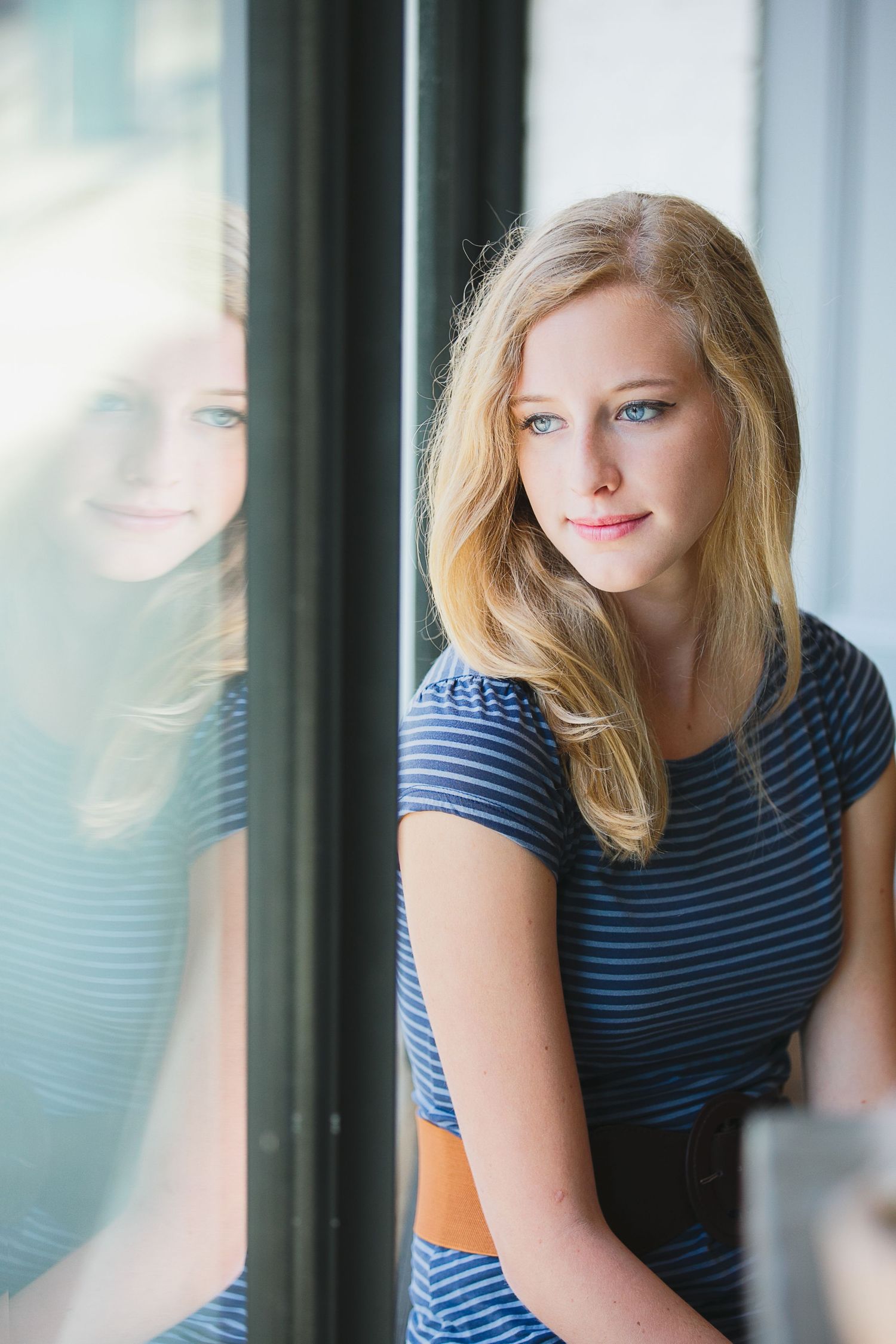 Young woman in striped shirt gazes thoughtfully by window with reflection.