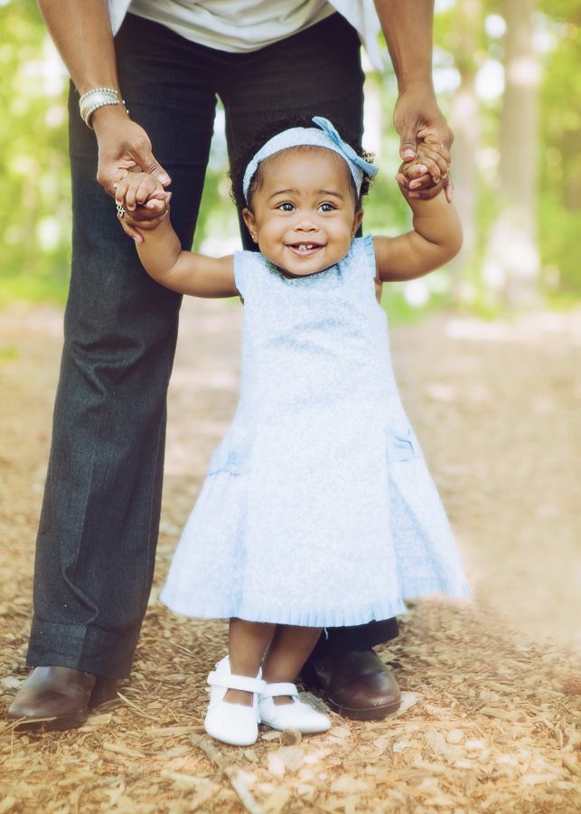 A small child in a white dress and hair bow takes their first steps while holding hands with an adult outdoors.