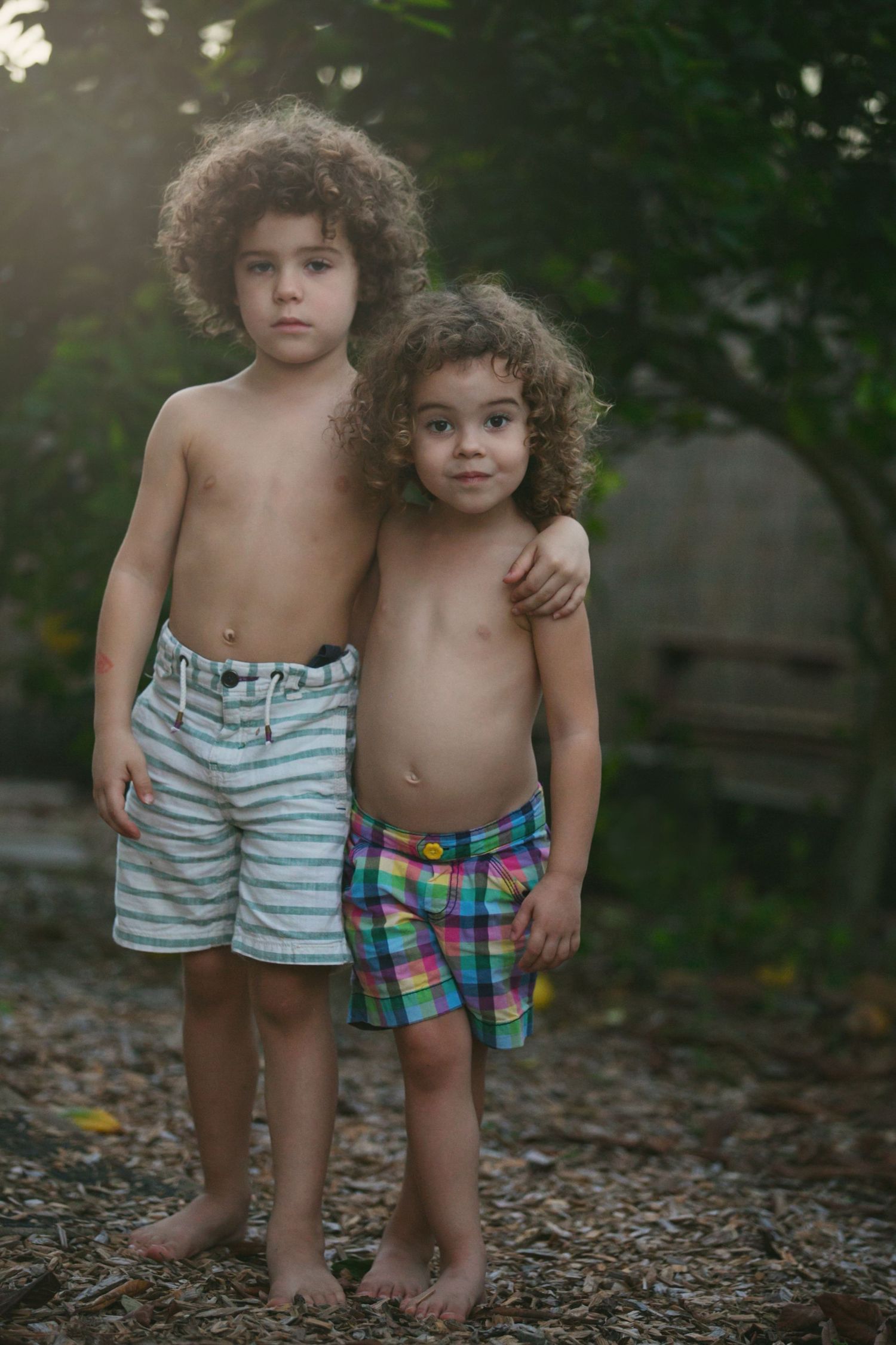 Two curly-haired siblings in swim shorts standing together on forest path at sunset.
