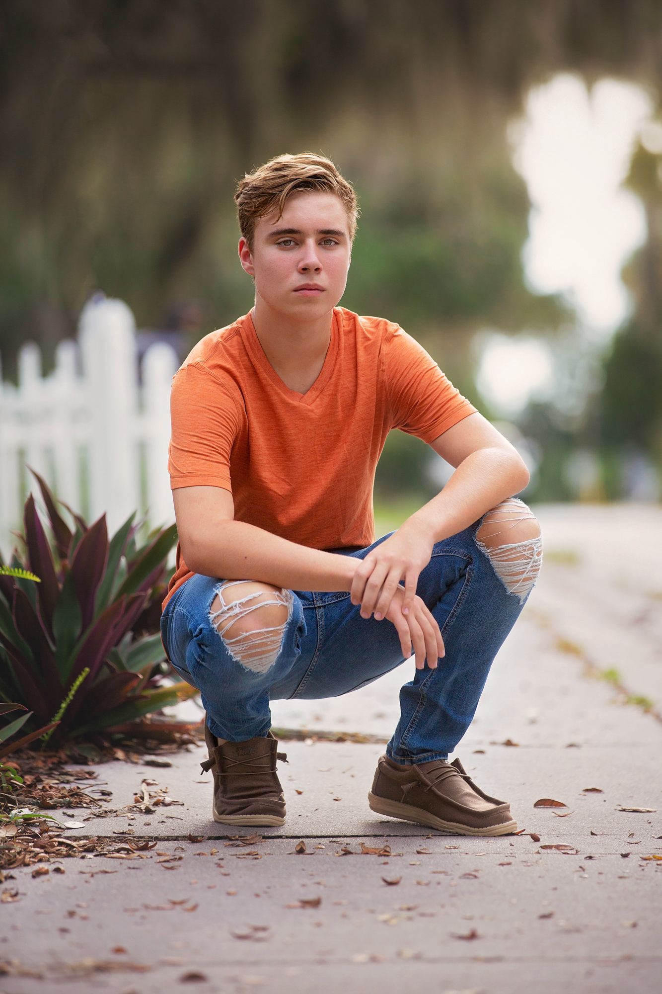 Fashion portrait of someone in orange shirt and ripped jeans crouching on sidewalk.