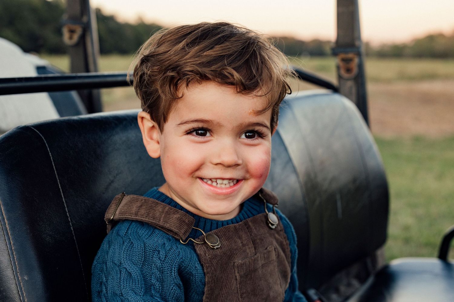 Child wearing blue shirt and brown vest sitting in vehicle seat outdoors.