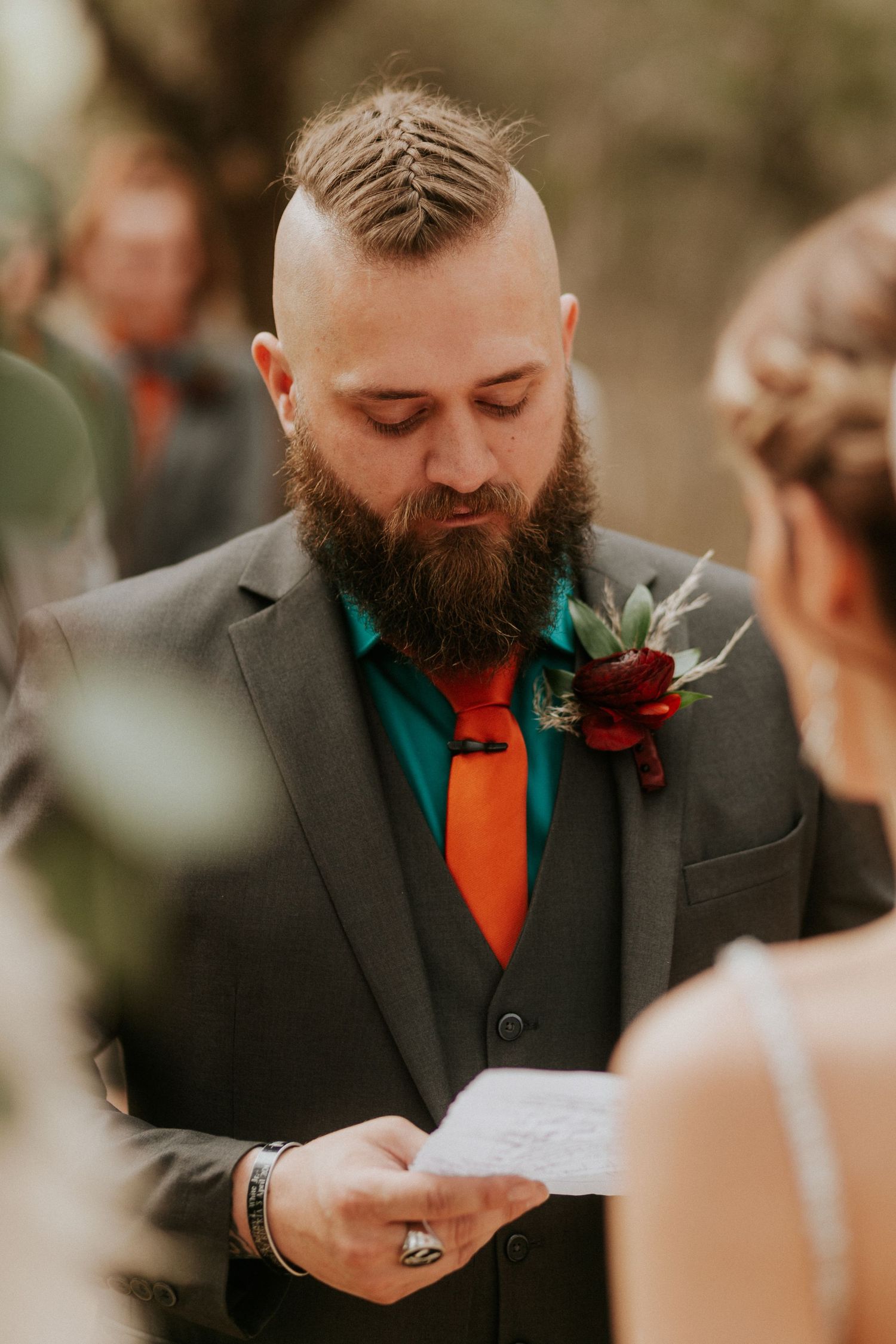 A groom with a mohawk and beard reads vows while wearing a gray suit with teal and orange accents.