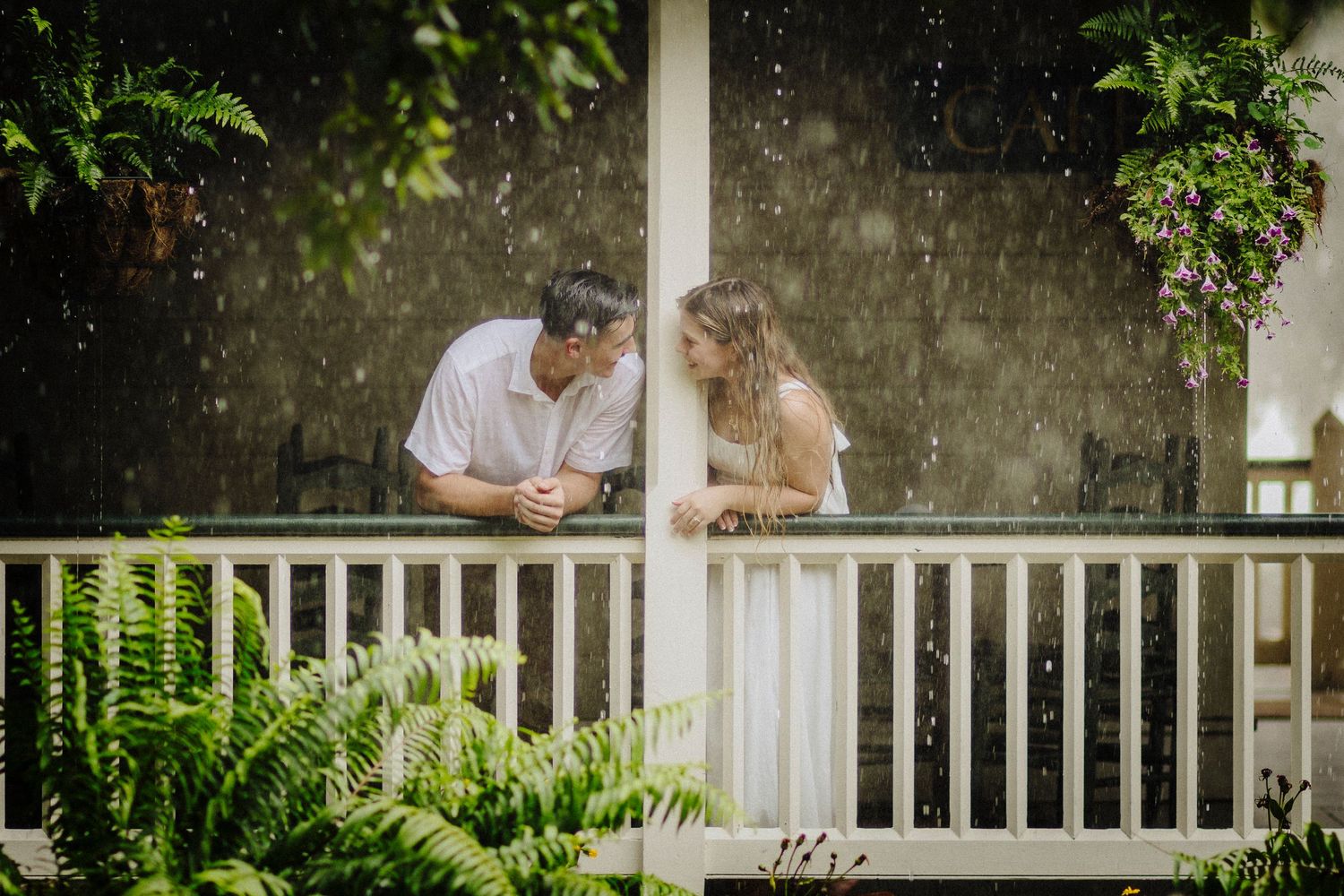 A couple shares an intimate moment on a white porch railing surrounded by lush green ferns and foliage.