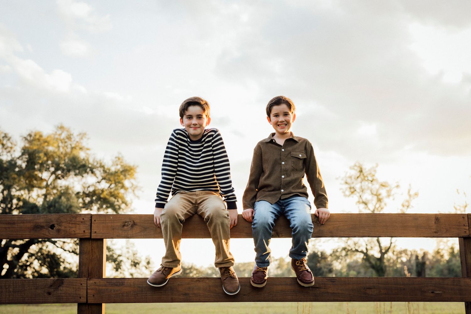 Two children sit side by side on a wooden fence at sunset.