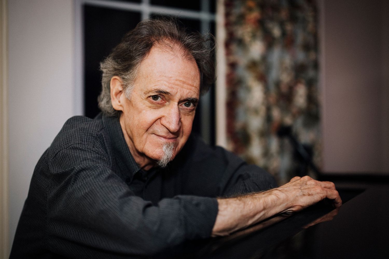 Portrait of an elderly man in dark clothing leaning thoughtfully against a surface in indoor lighting.