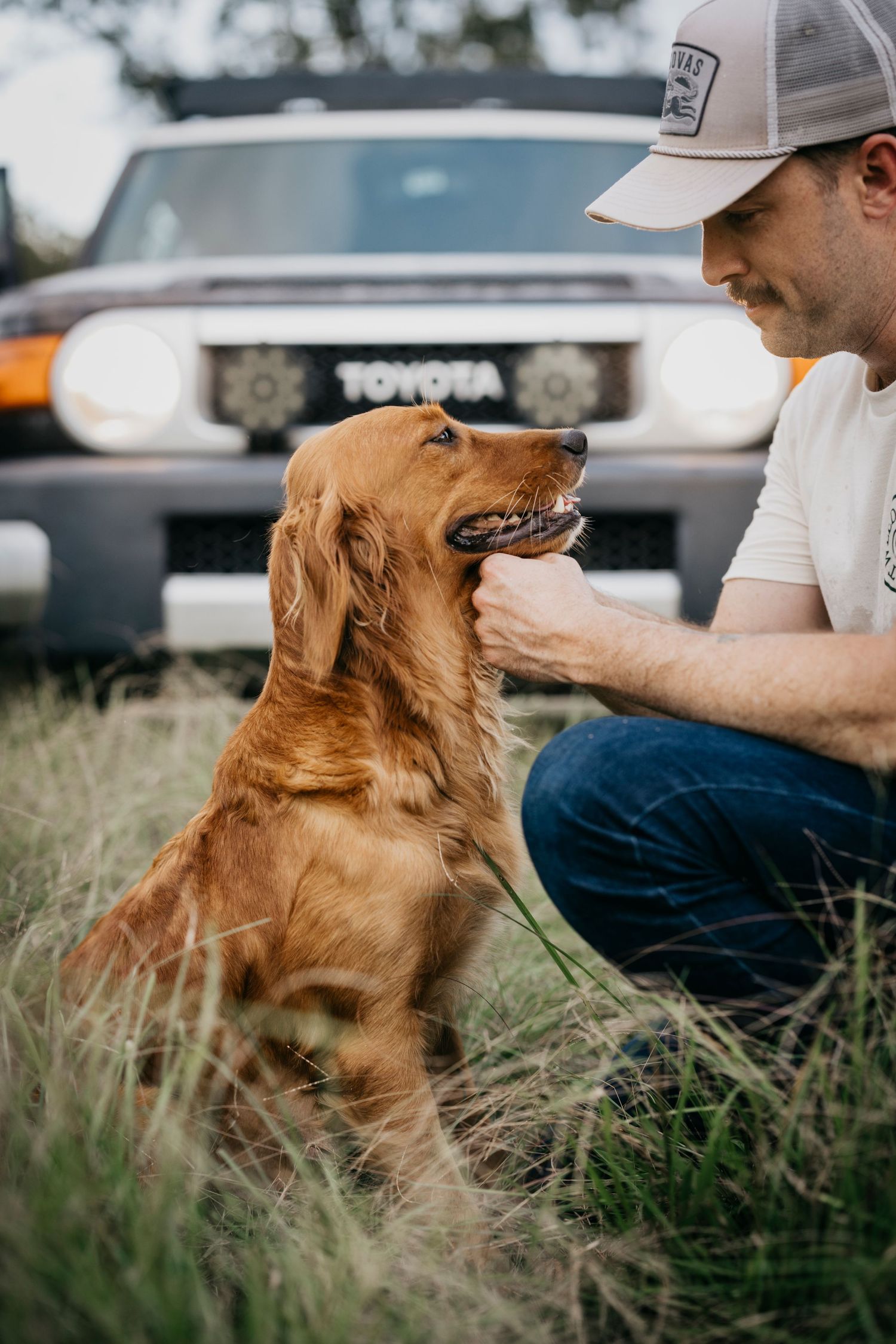 Golden retriever and owner share a moment beside Toyota truck in tall grass.