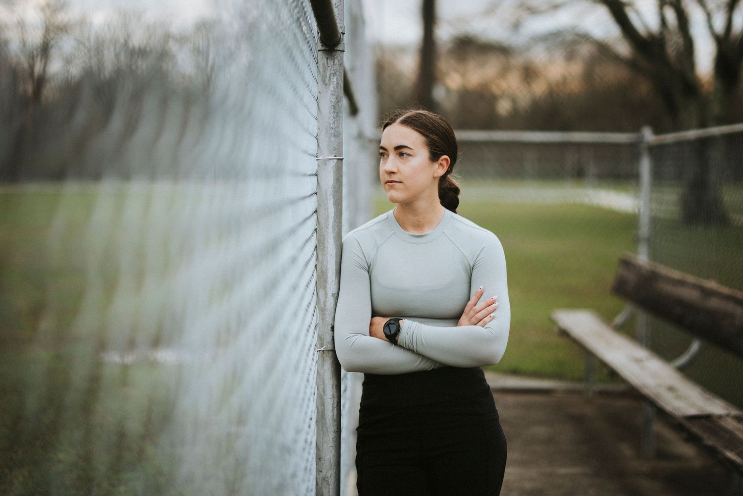 Person in gray sweater stands with arms crossed by white mesh fence at outdoor sports field.