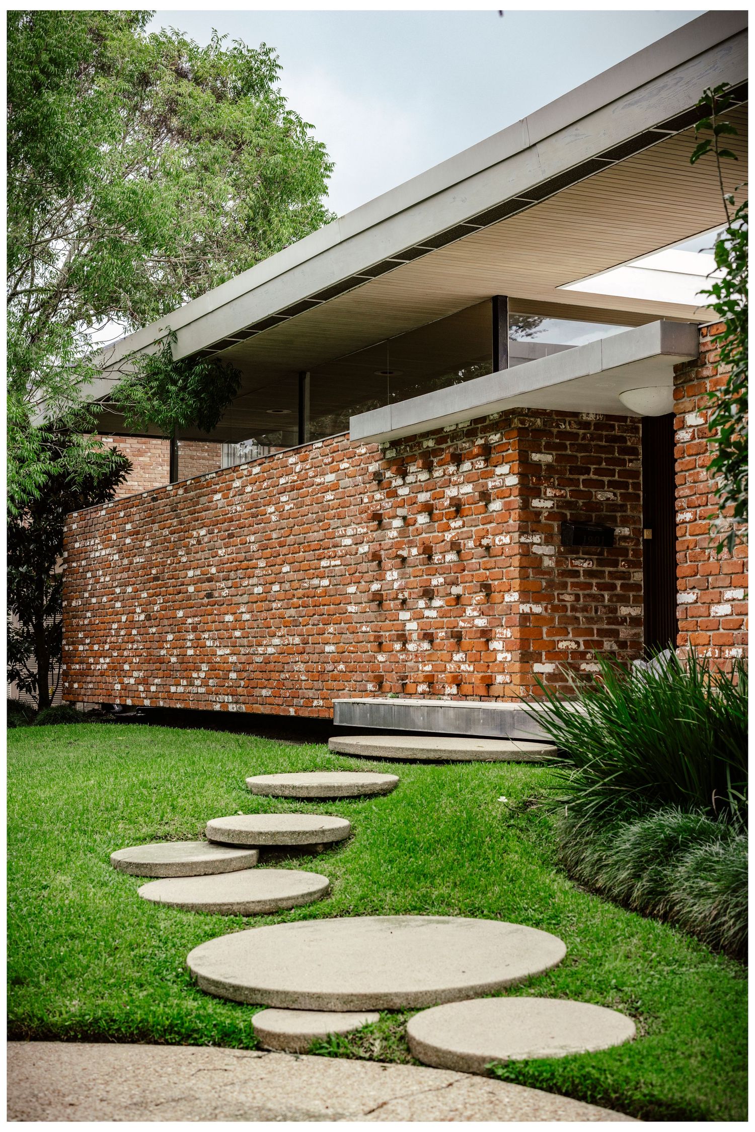 Modern architectural design features curved stepping stones leading to a brick wall with geometric cutouts and overhanging roof.