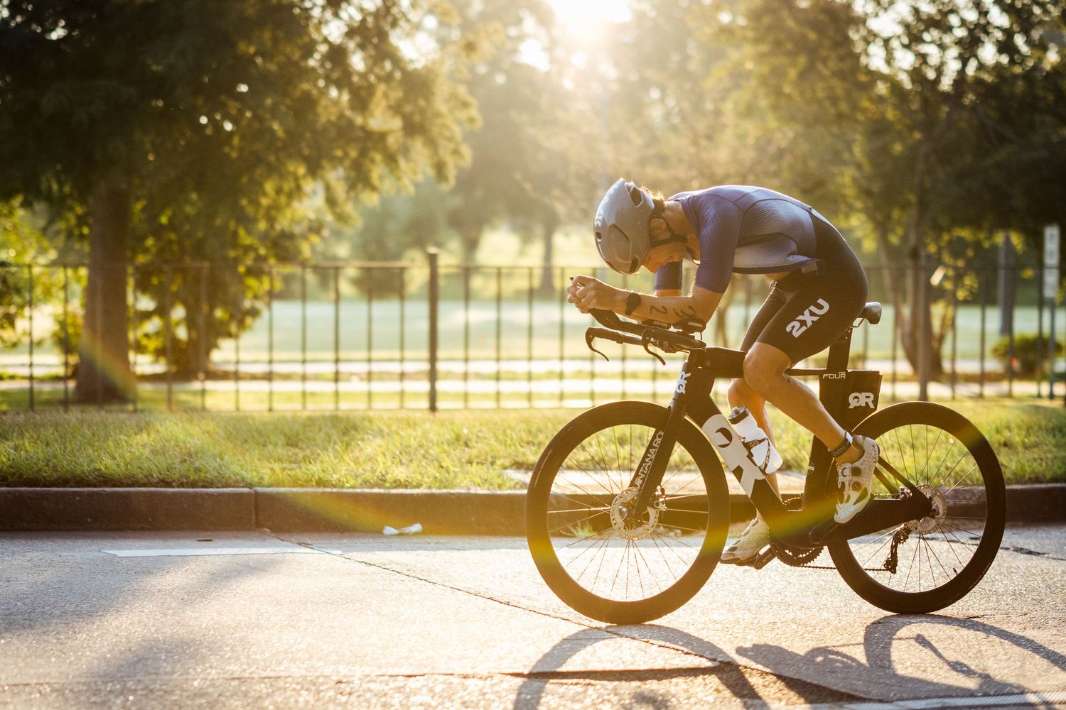 Silhouetted cyclist rides time trial bike at sunset through park setting.