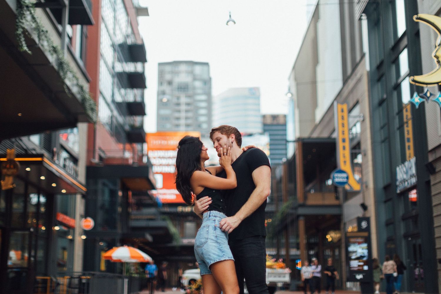 A couple shares an intimate moment on a city street with urban buildings and orange umbrellas in the background.