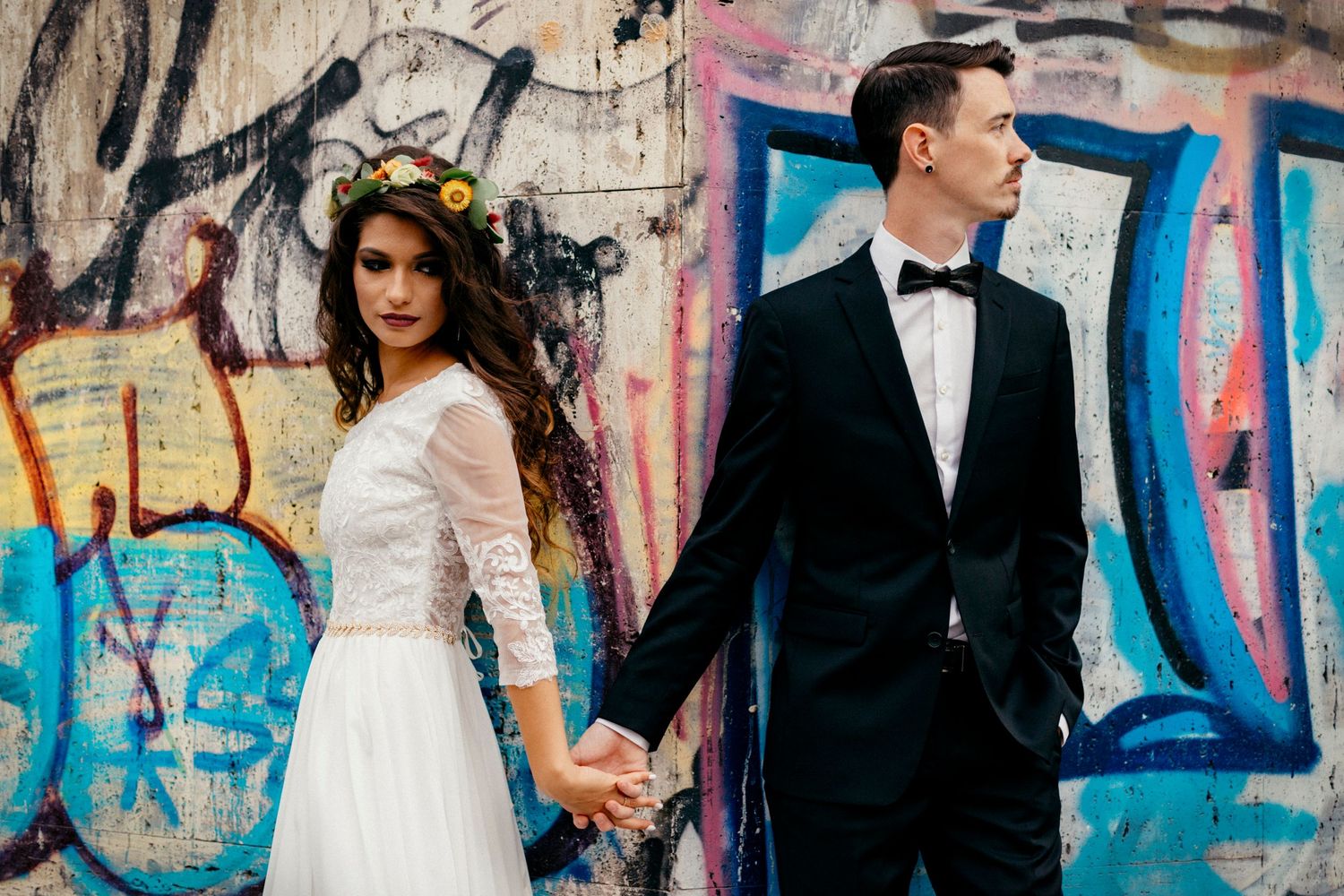 Newlyweds pose against colorful graffiti wall in wedding attire holding hands.