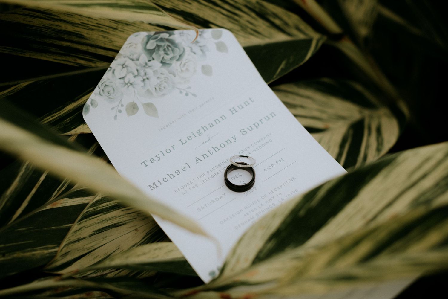 Wedding invitation and ring displayed on tropical palm leaves with soft lighting.