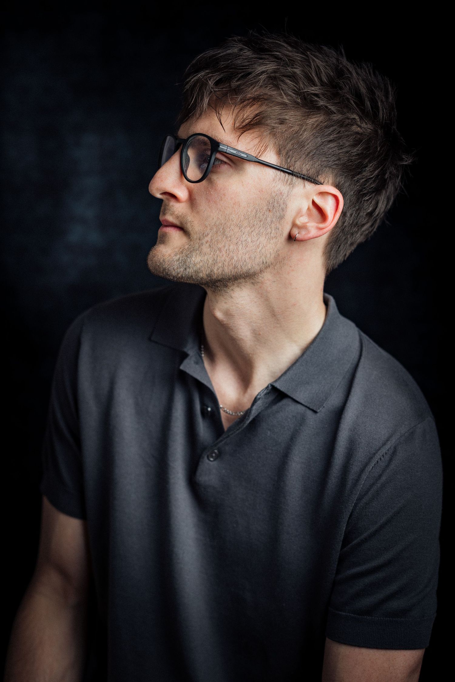 Dramatic side profile portrait in grey polo and glasses against dark background.