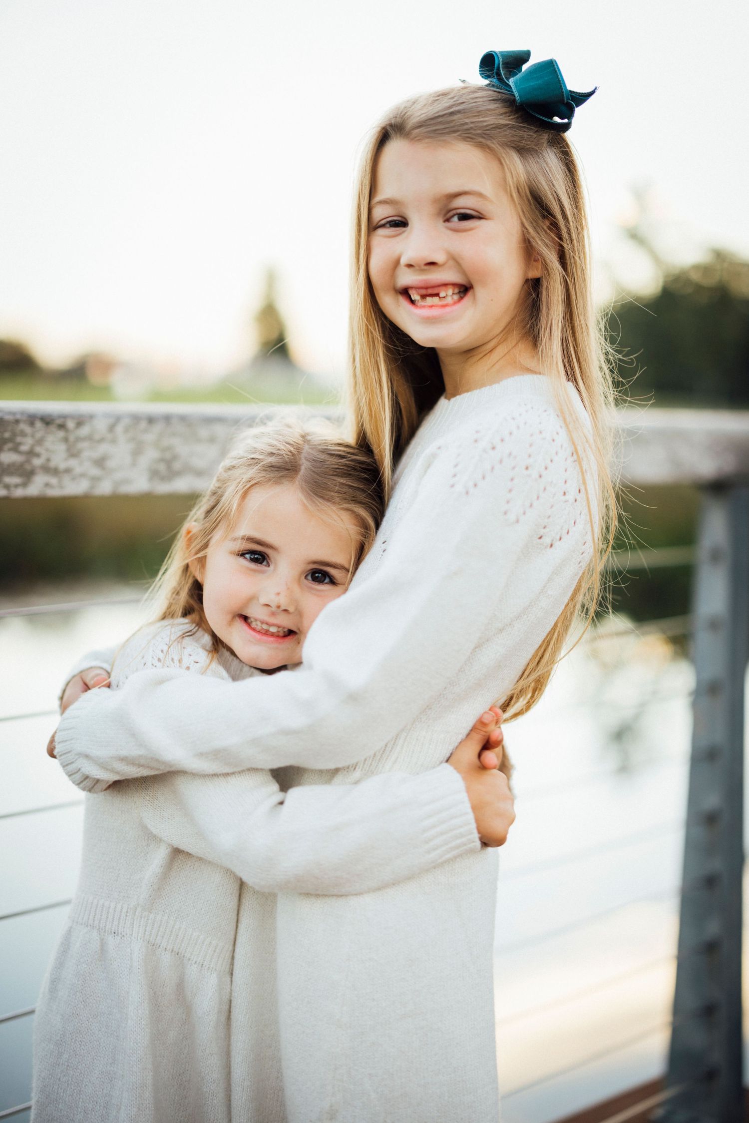 Two sisters in white sweaters embrace while smiling at the camera.