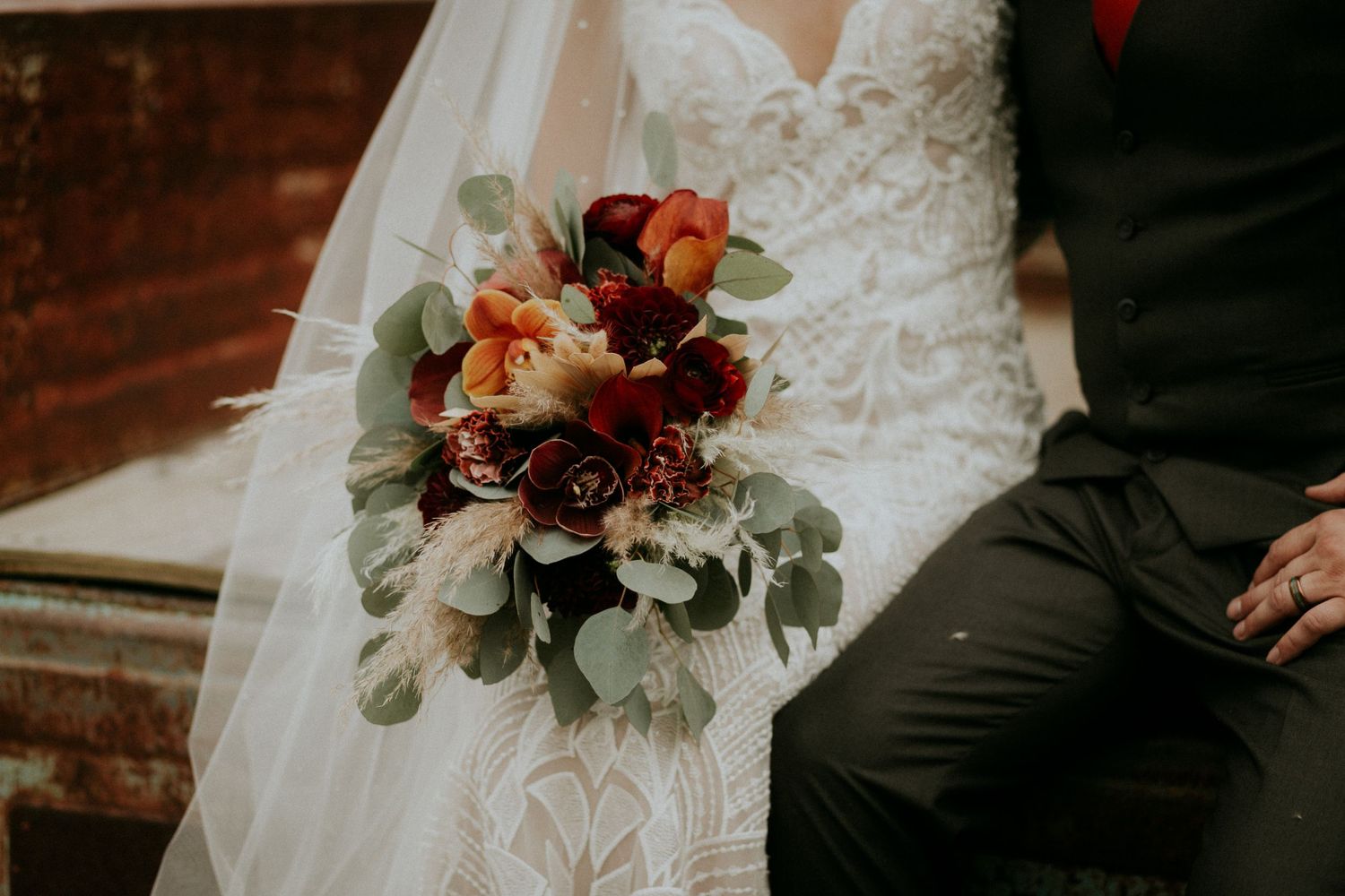 Wedding bouquet detail with burgundy flowers and eucalyptus on lace dress.