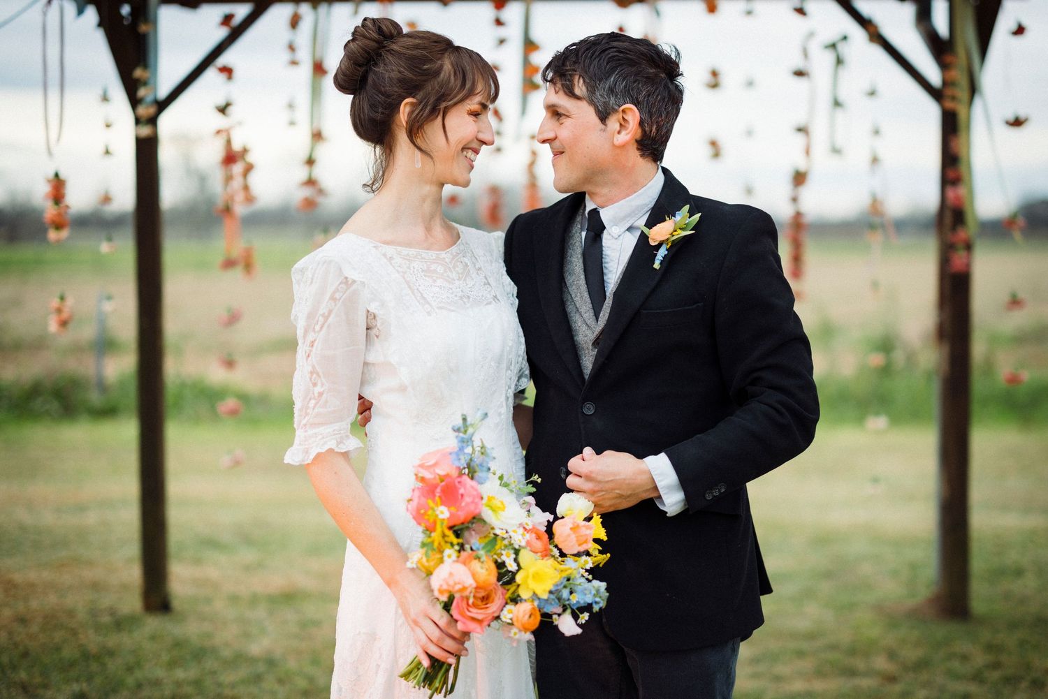 A couple in wedding attire shares a romantic moment among trees with hanging orange floral decorations.