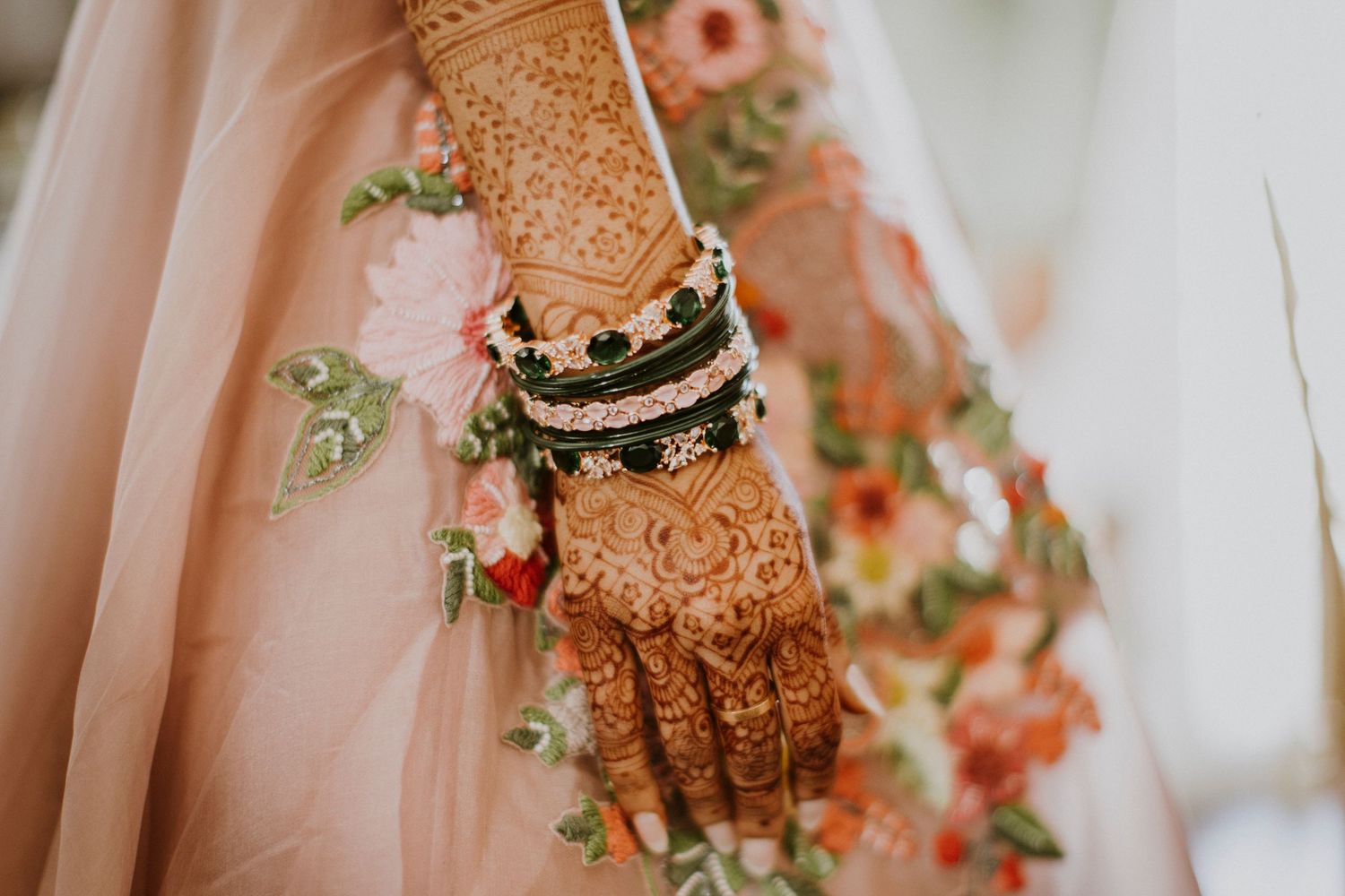 Intricate henna design adorns a hand wearing gold bangles and jewelry against a soft pink fabric background.
