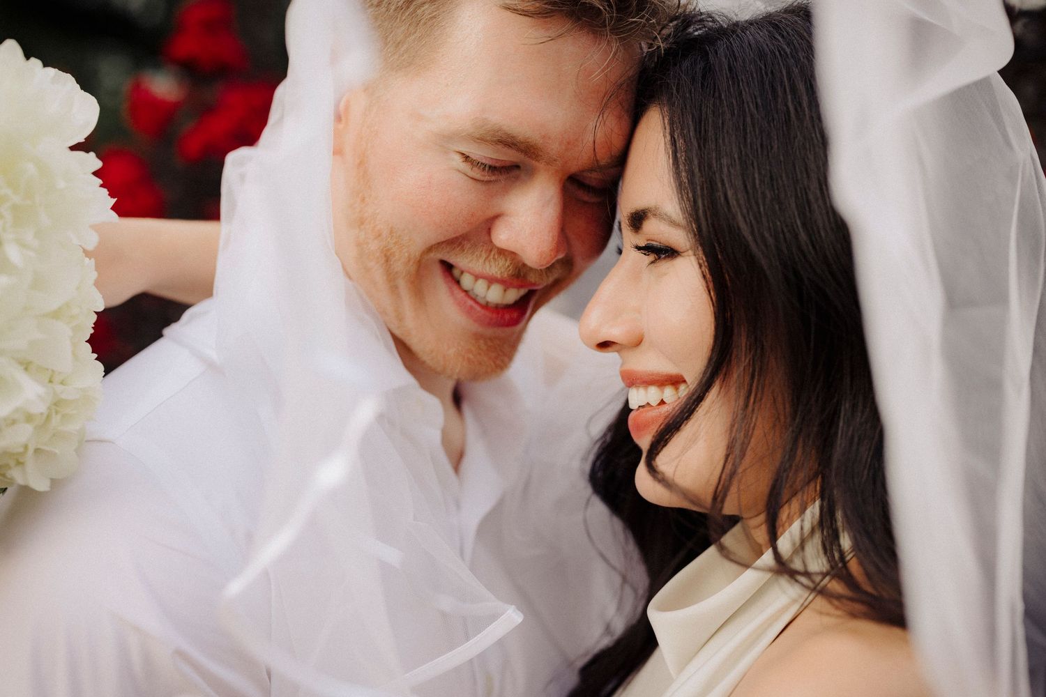 A joyful couple shares an intimate moment under a white veil with red and white flowers in the background.