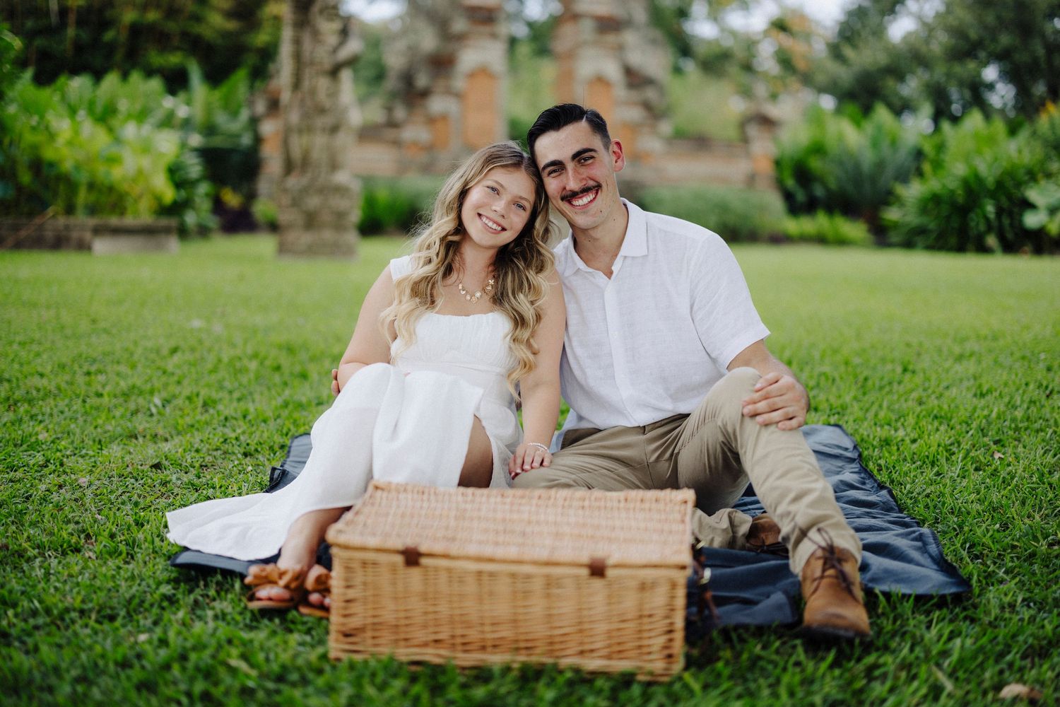 A couple enjoys a romantic picnic date on lush green grass with a wicker basket and blanket on a sunny day.