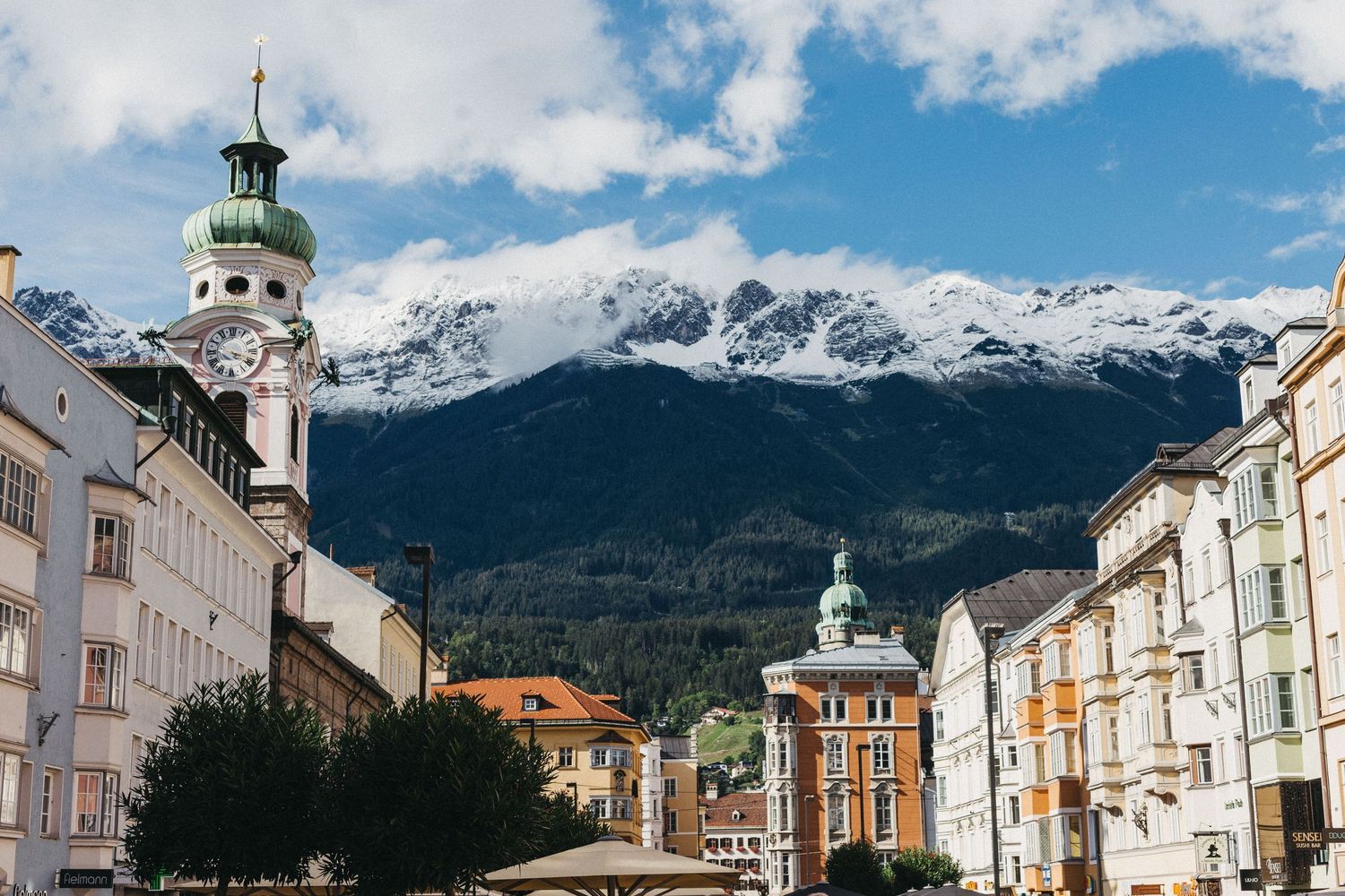 Historic European street with church spire and snow-capped mountains in background.