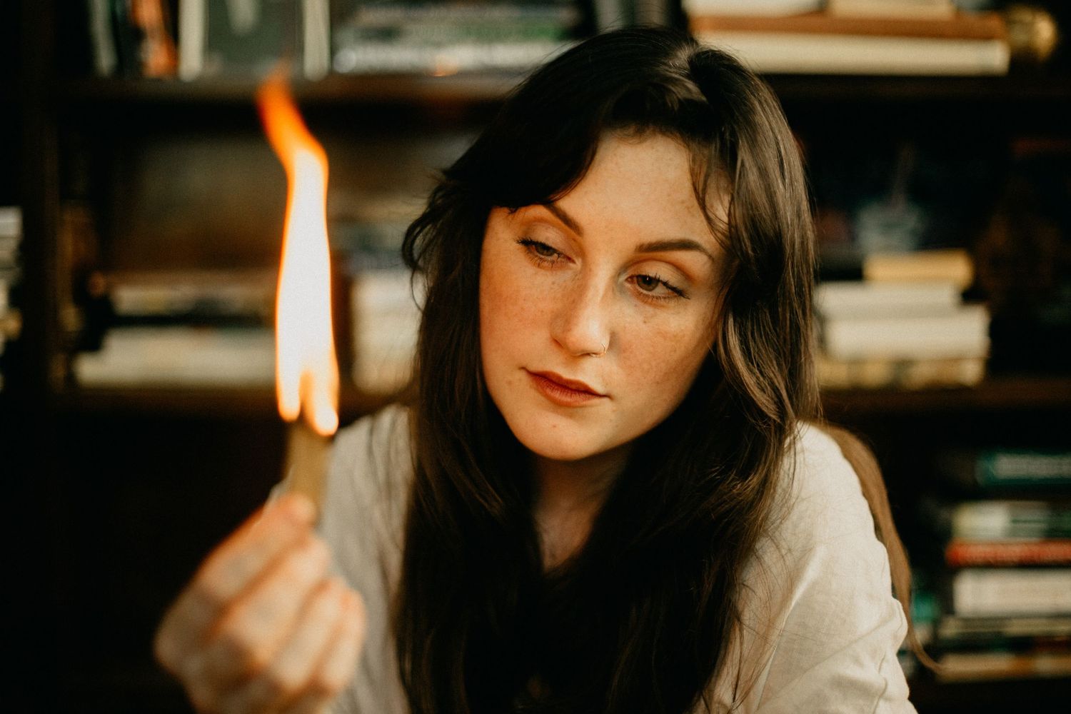 A close-up portrait showing someone holding a burning match in a dimly lit room with bookshelves in the background.