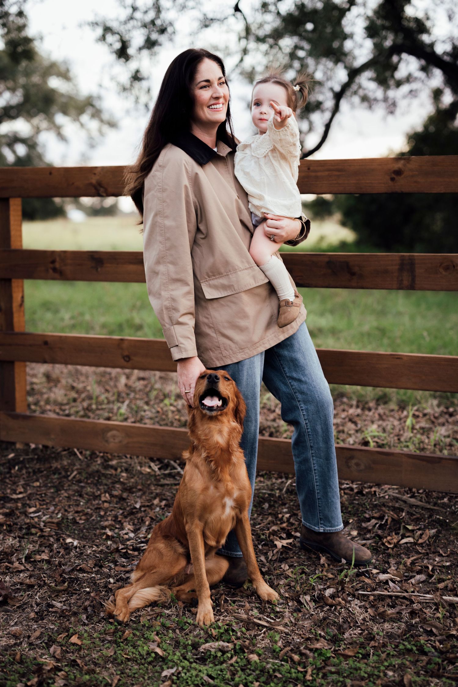 Woman and child standing by wooden fence with brown dog sitting obediently beside them.