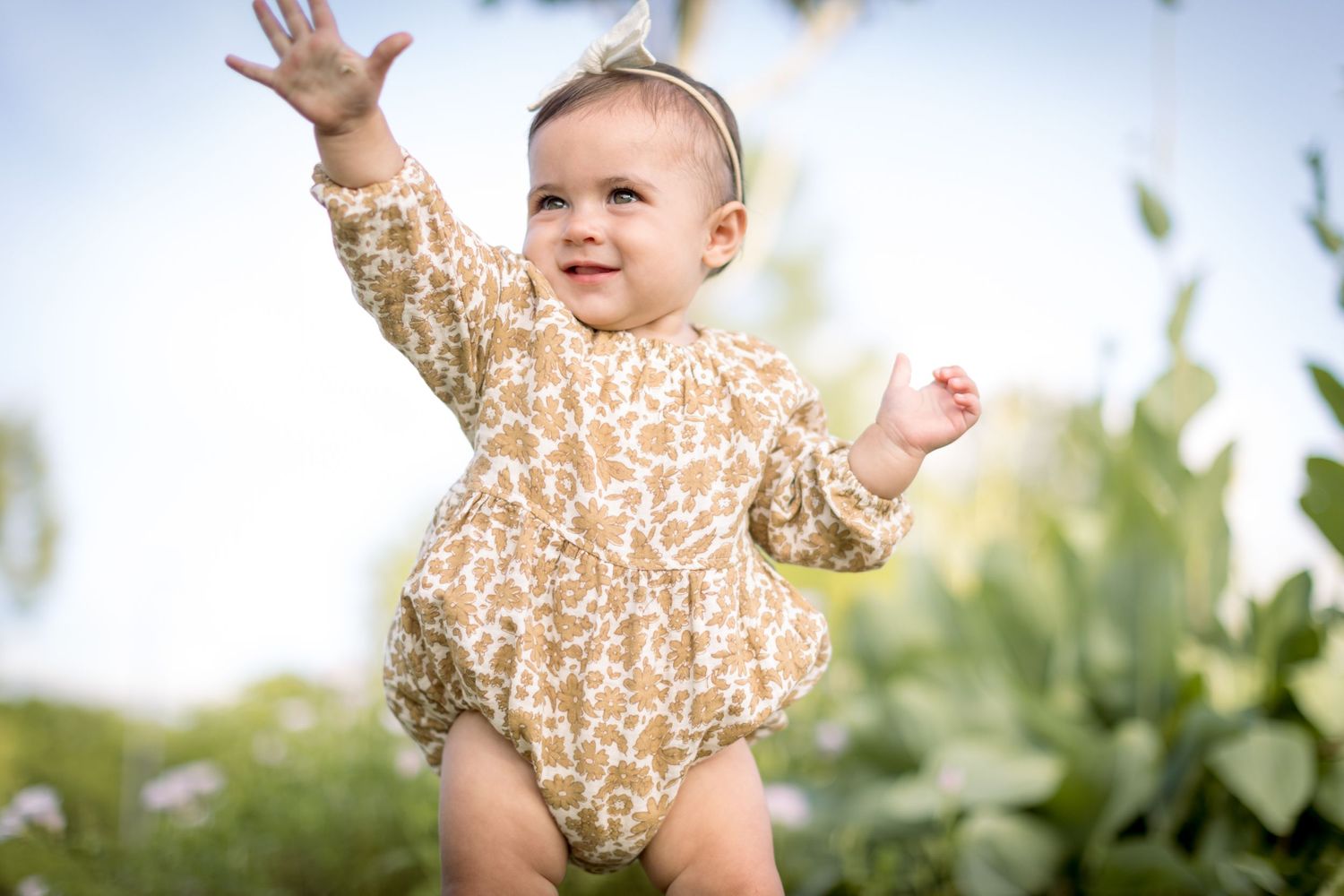 Happy baby in floral romper reaching up during outdoor photoshoot.