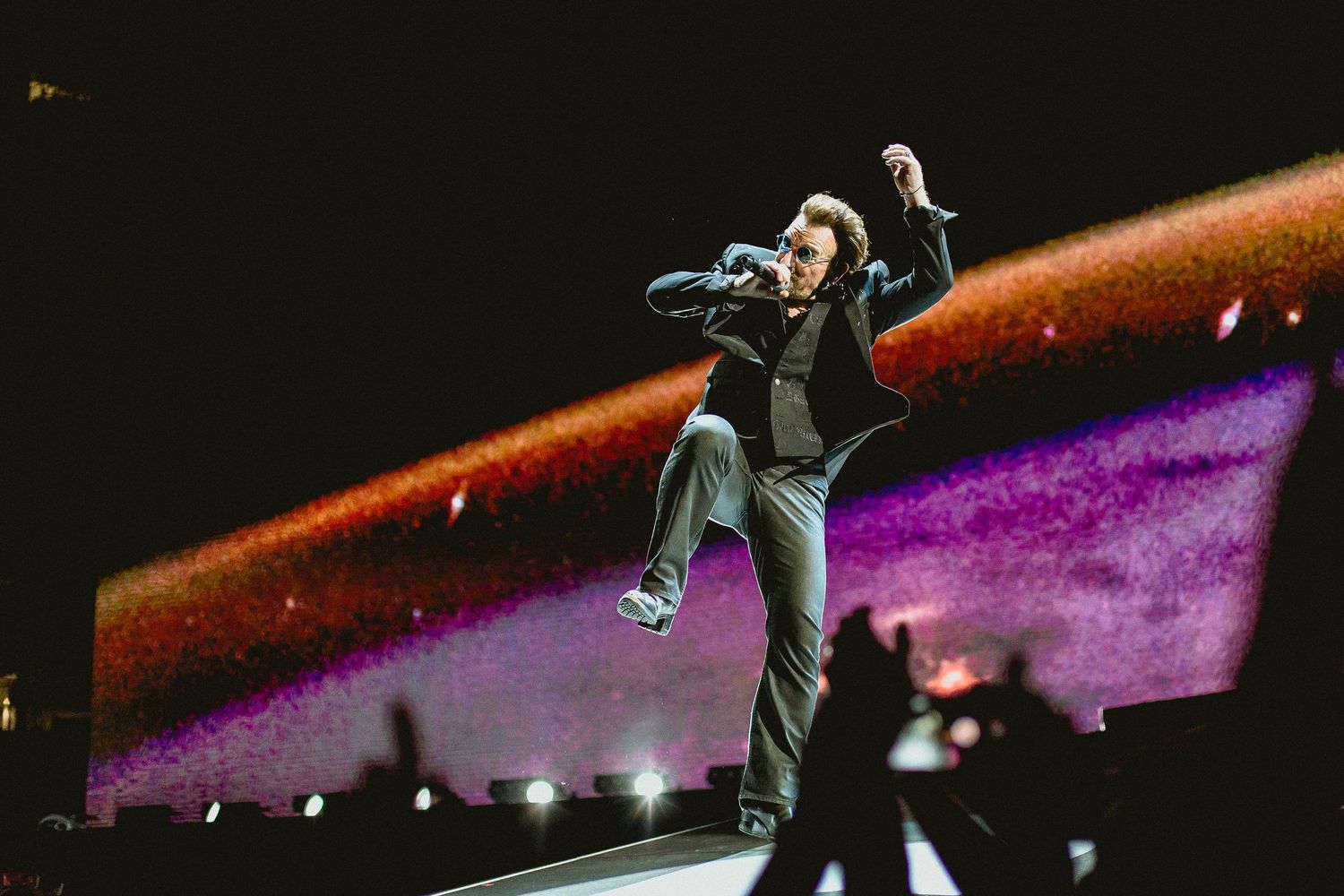 A concert performer leaps energetically on stage with purple and orange lighting effects.