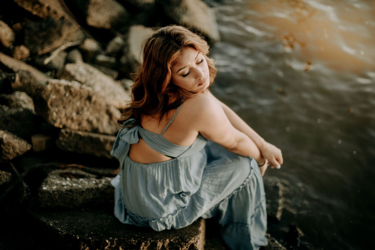 Portrait of someone in a blue dress sitting on coastal rocks at sunset.