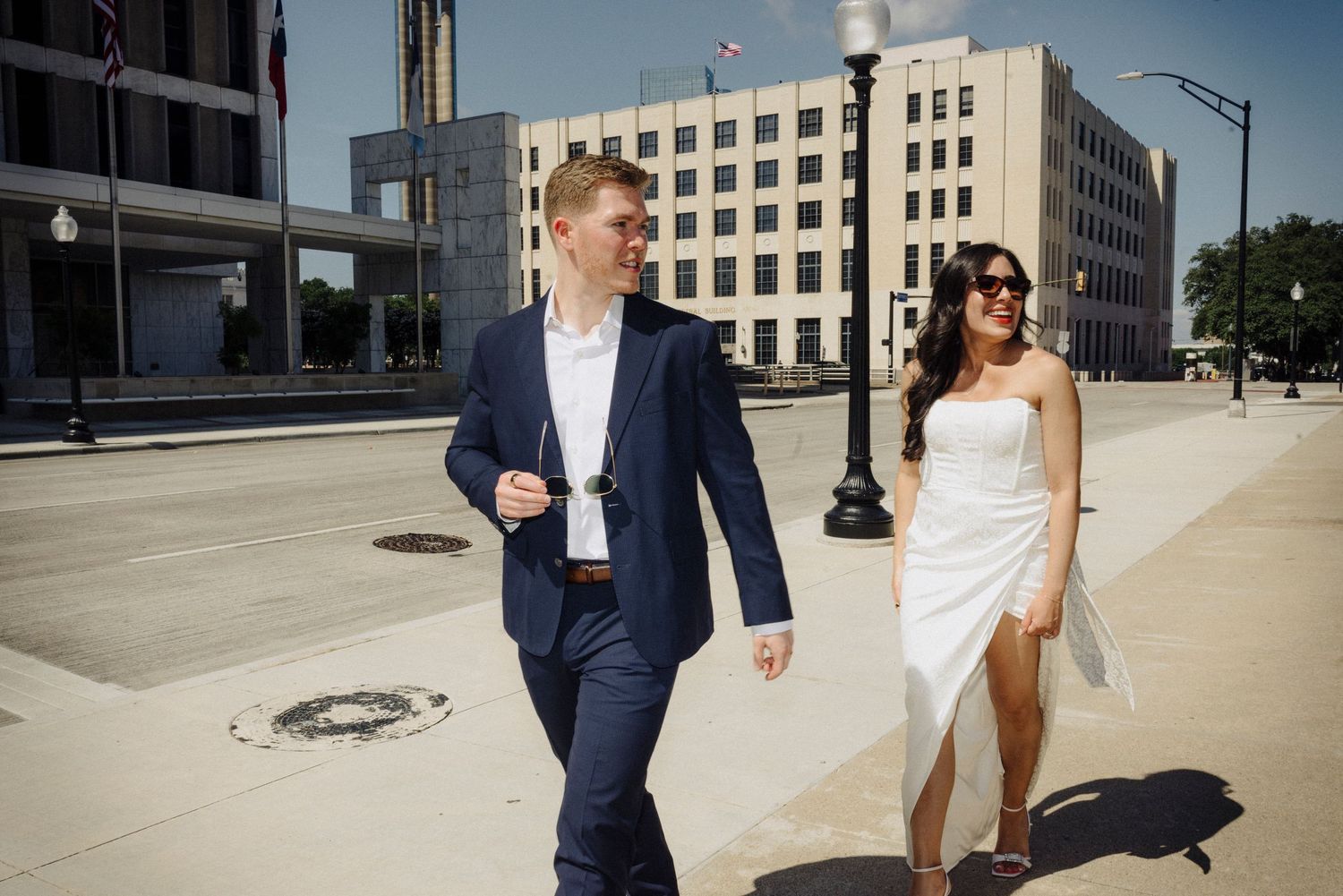 A couple in wedding attire walks along a city sidewalk with tall buildings in the background on a sunny day.