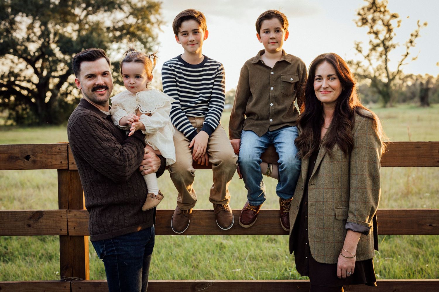 A family sits together on a wooden fence at sunset in a field.