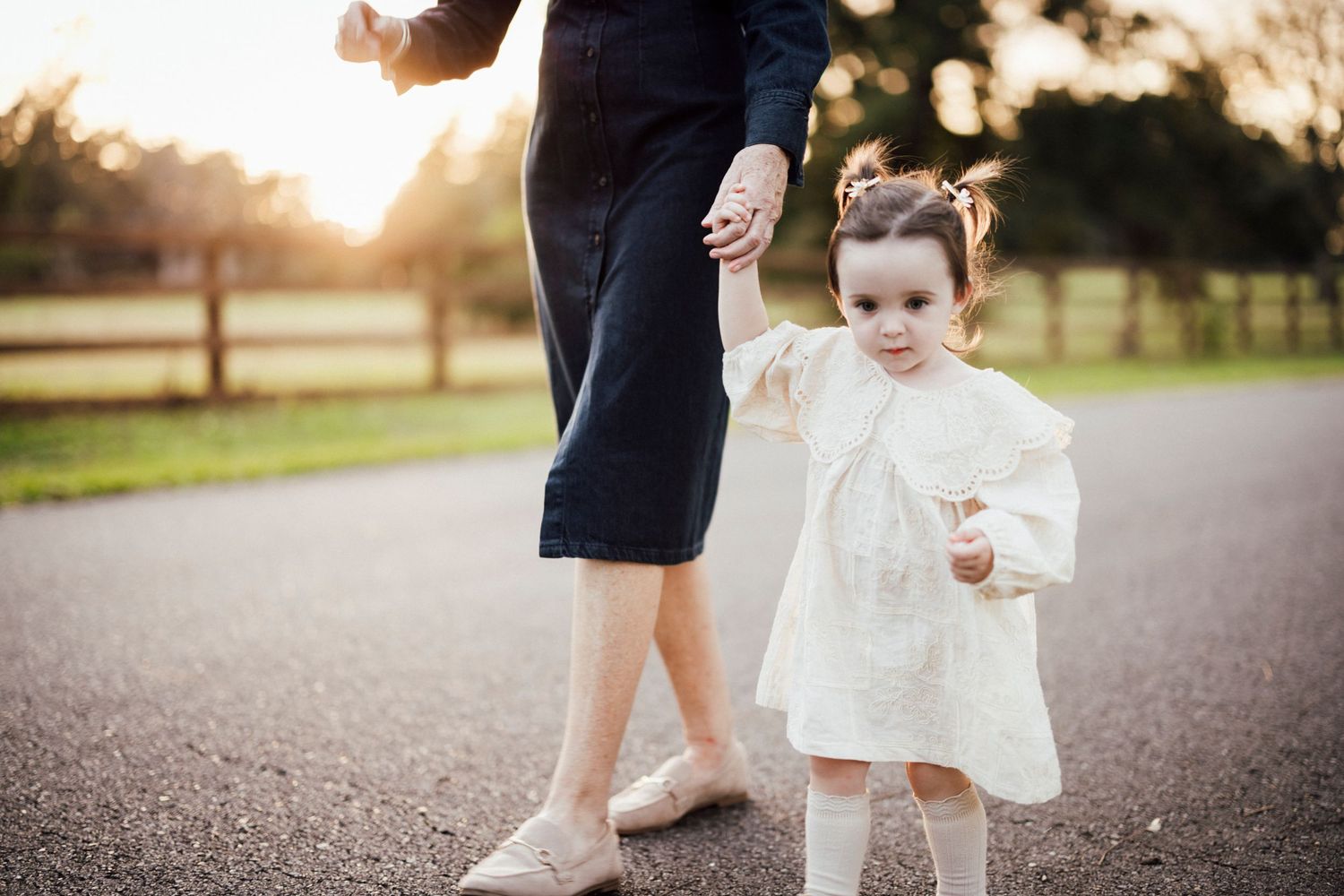 Small child in white dress holding hands with parent walking on path at sunset.