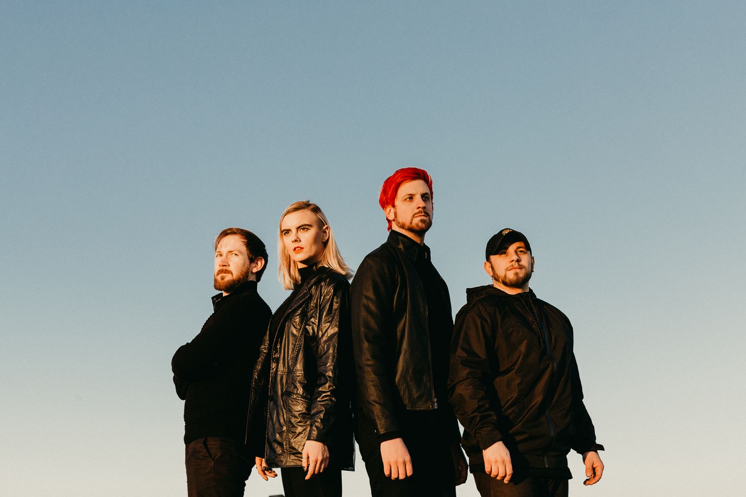 Rock band members in dark clothing pose against clear blue sky.