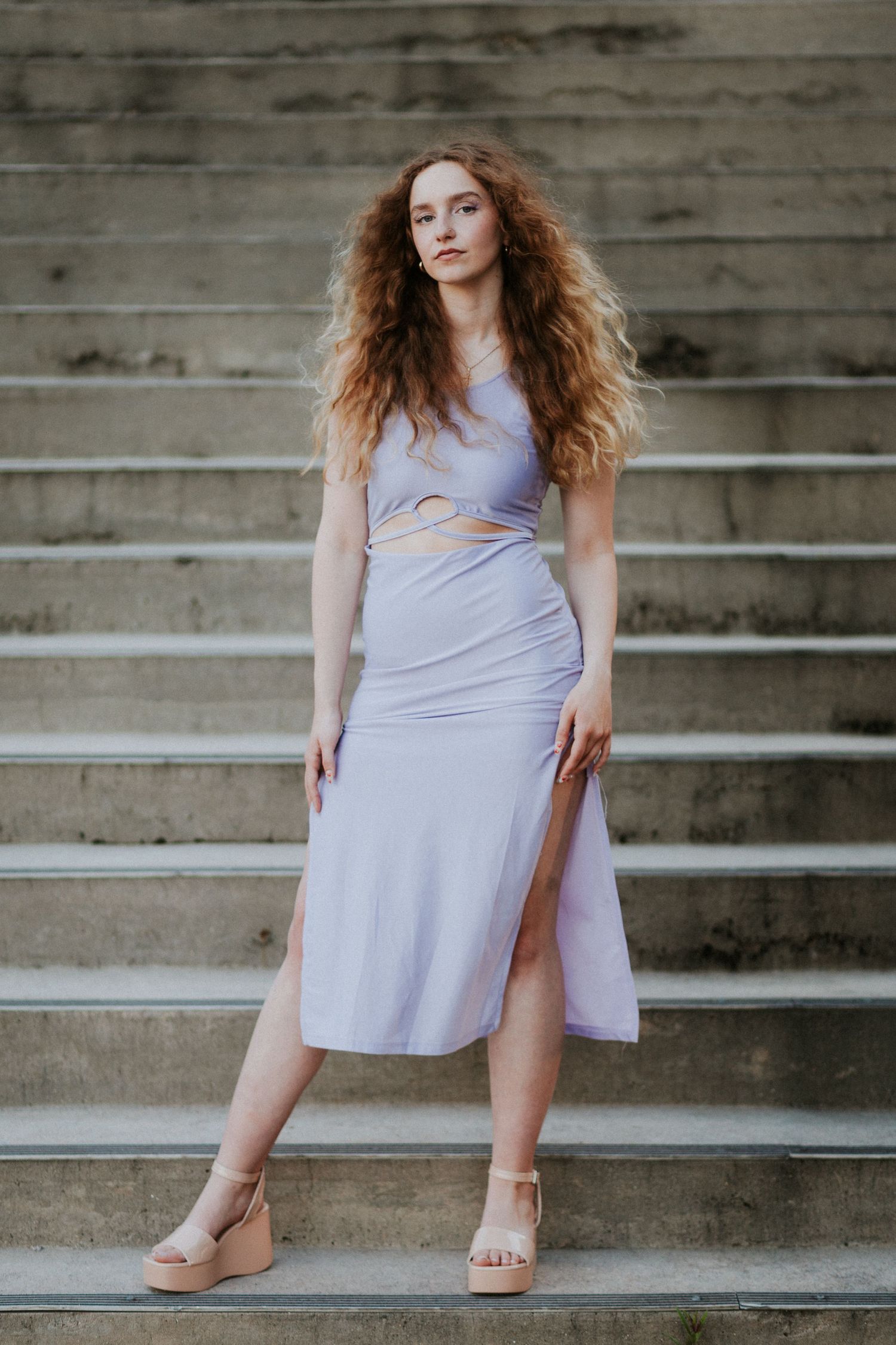 A woman with flowing red hair poses on concrete steps wearing a lilac cutout dress.