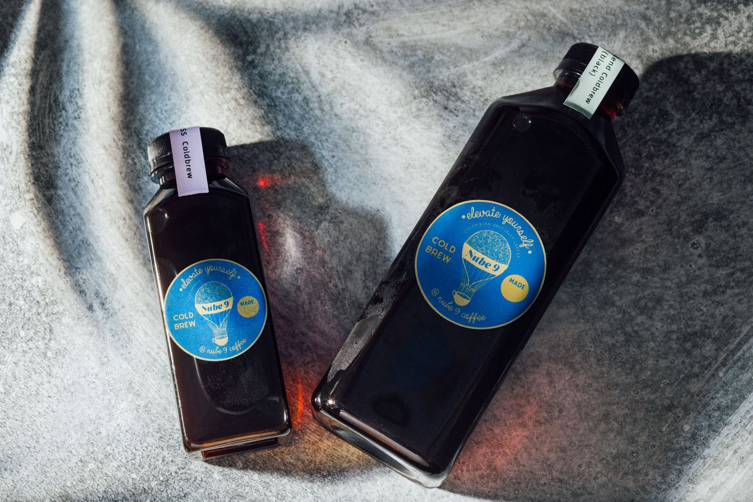 Two dark glass bottles with blue circular labels photographed on reflective surface.