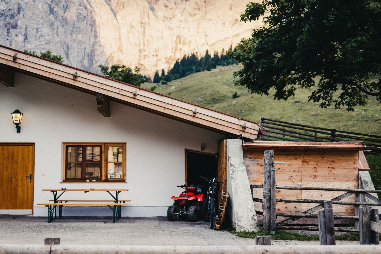 Rustic alpine cabin with wooden benches and a red ATV parked outside against mountain backdrop.