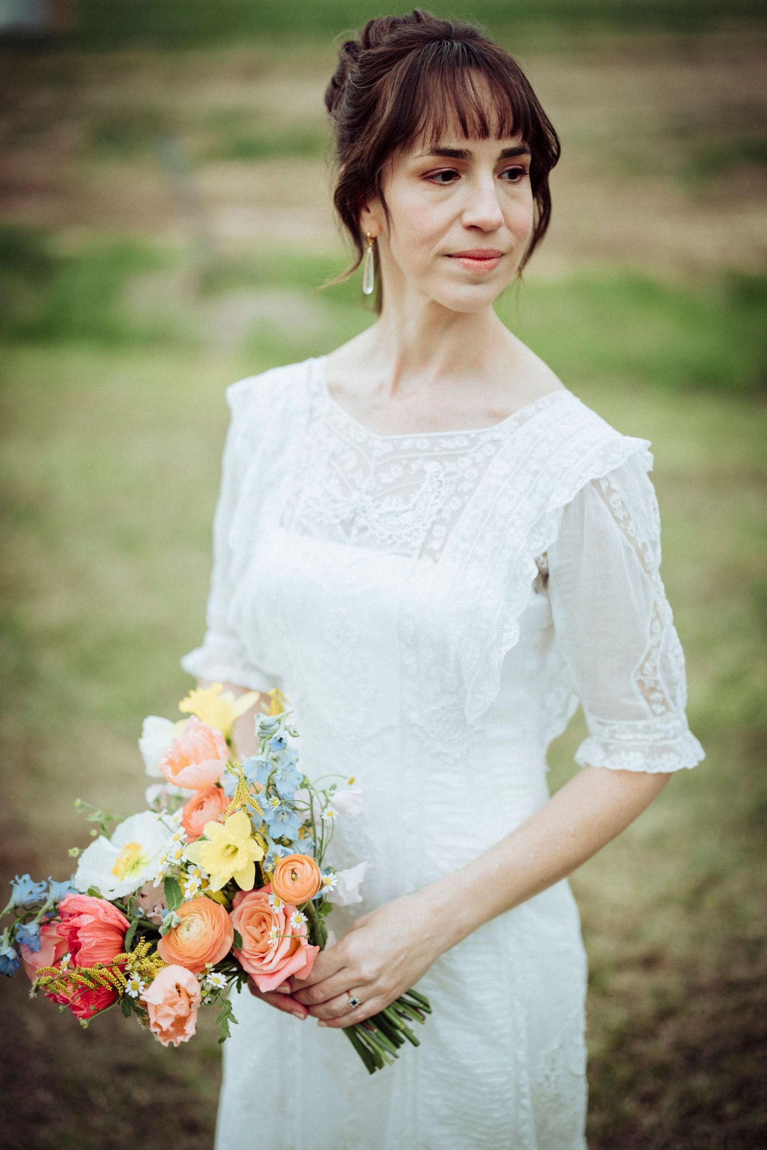 A bride in a vintage-style lace dress holds a vibrant bouquet of pink and peach roses.