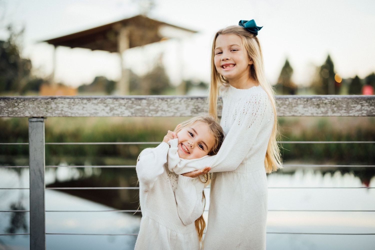 Two siblings in white sweaters share a joyful hug by a railing.