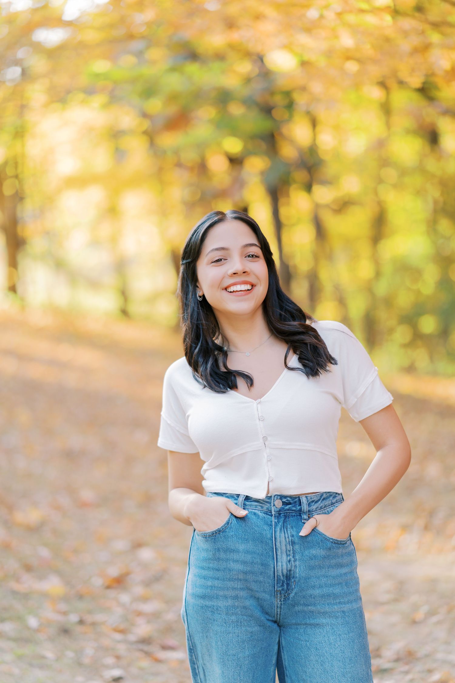 Person in white t-shirt and blue jeans poses outdoors with autumn foliage and golden sunlight in background.