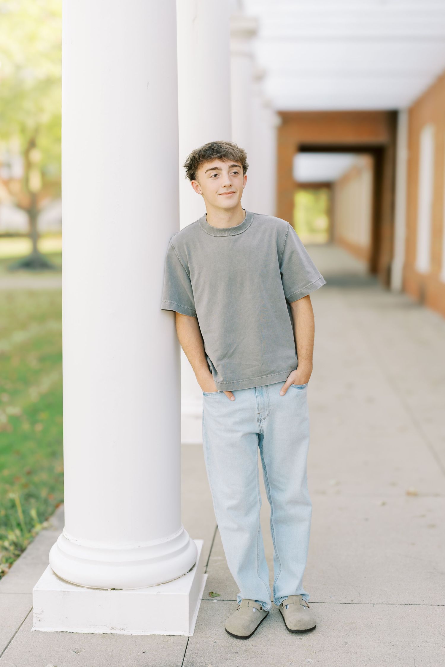 A person in a gray t-shirt and light jeans stands casually leaning against a white column in an outdoor walkway.