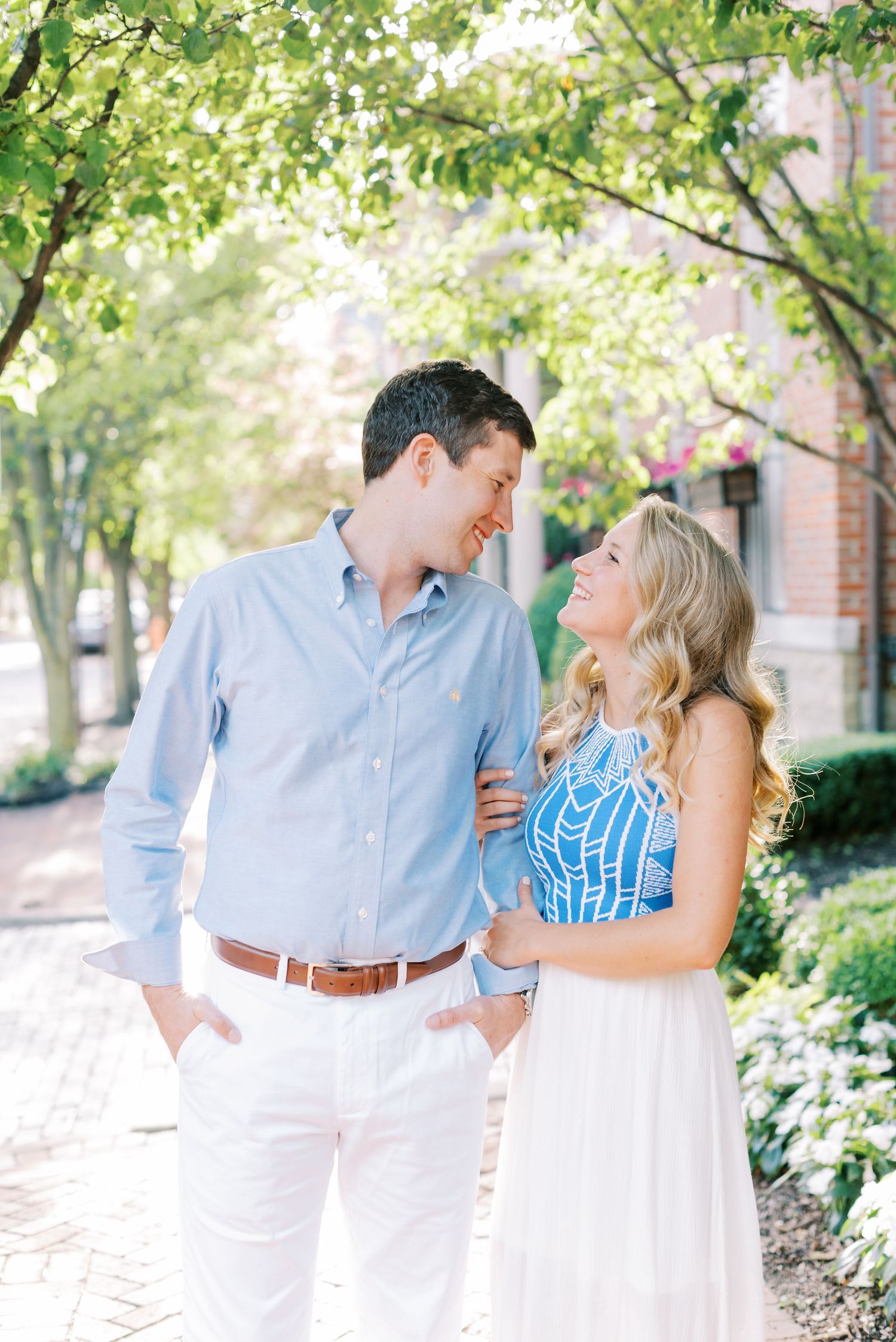 A couple in light blue and white outfits share joyful moments together in a garden setting with green trees and brick walls.