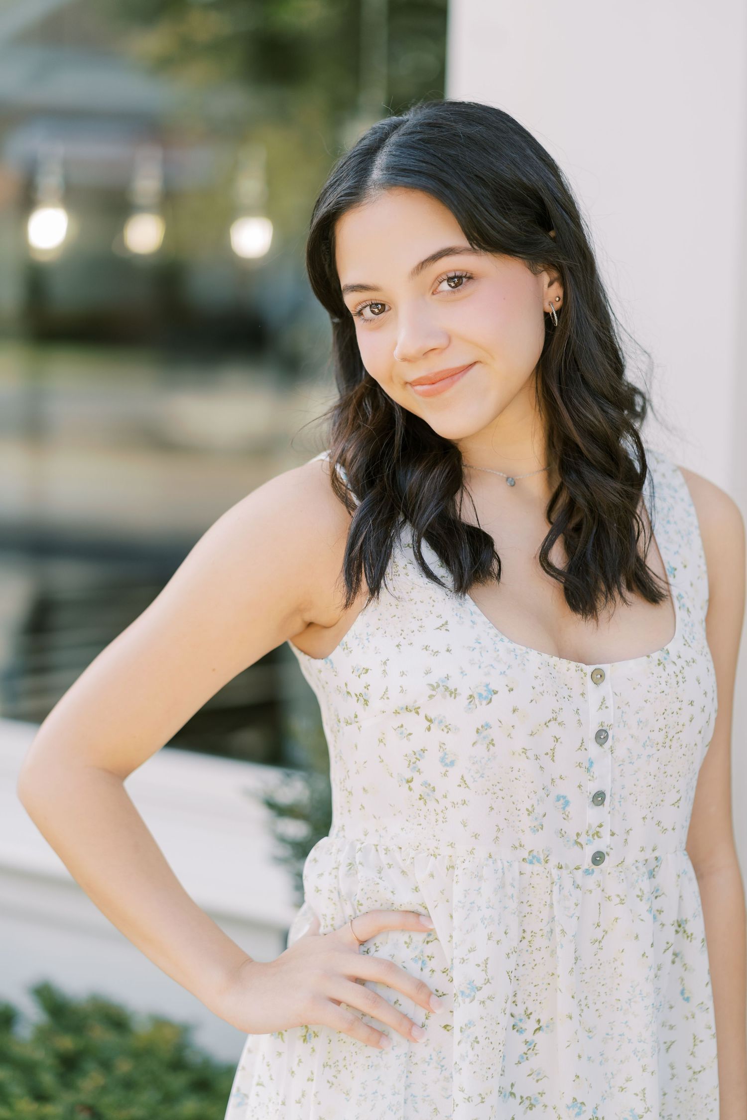A person in a white lace sundress poses outdoors with string lights and greenery in the background.