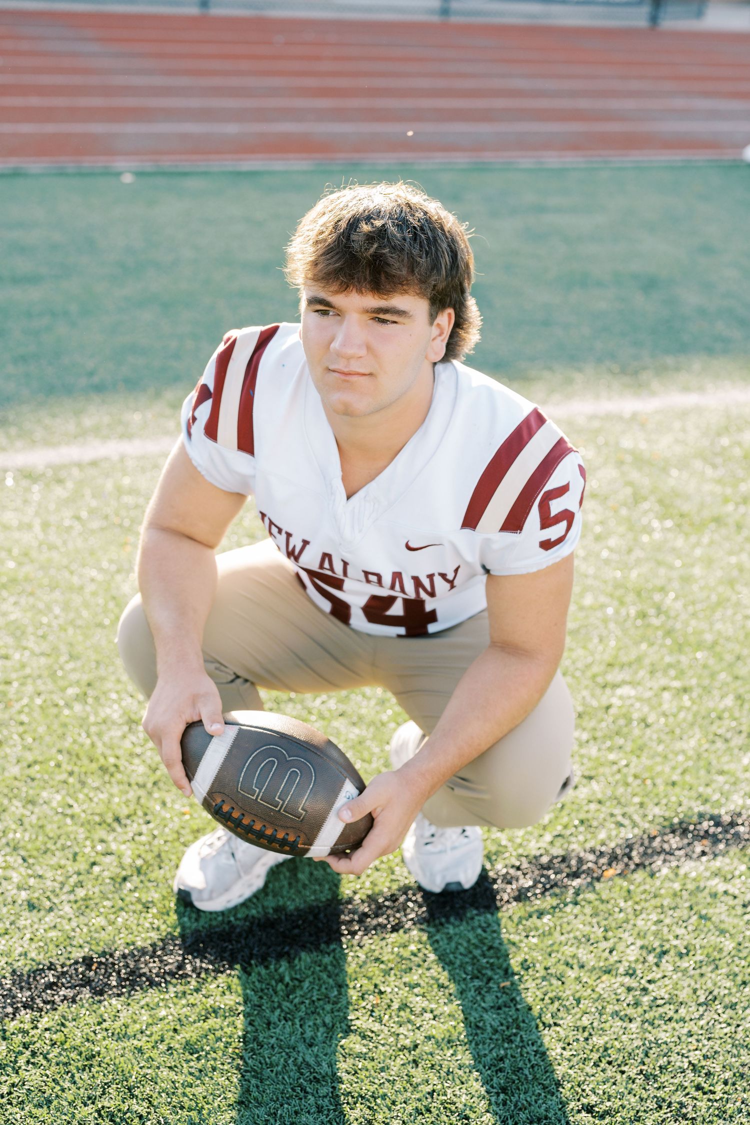 Football player in white and maroon uniform crouches on grass field while holding ball during practice.